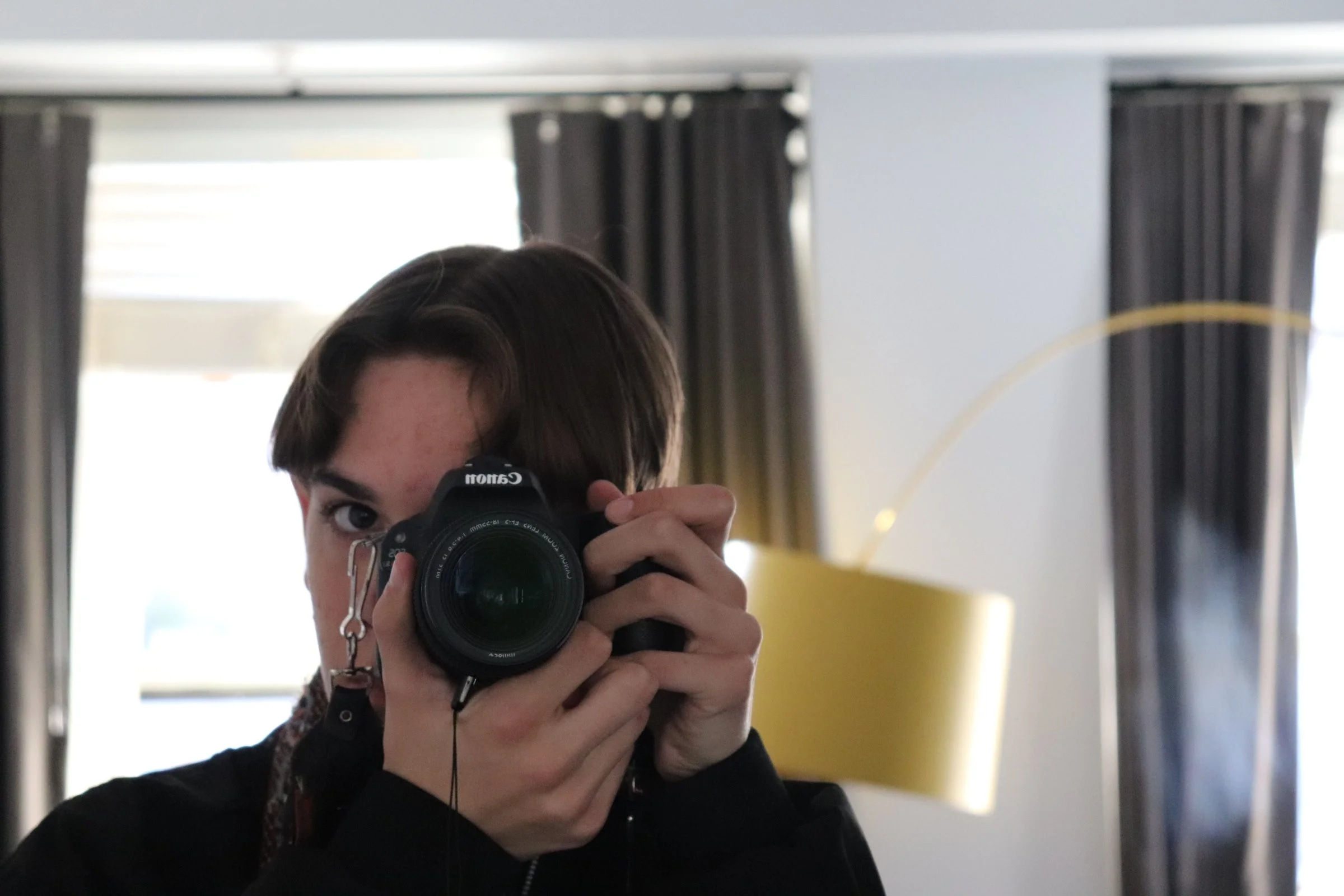 Person with short brown hair taking a photo with a Canon camera, partially obscuring their face, in a room with dark curtains and a yellow lamp
