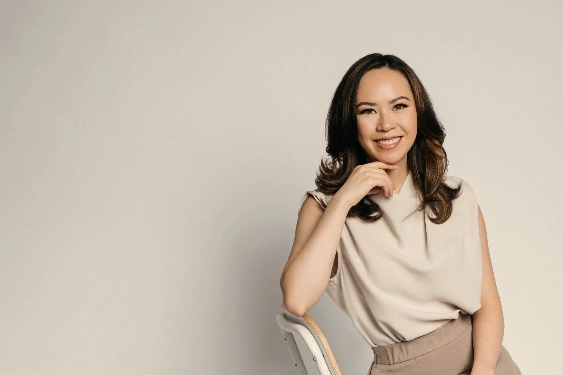 A young woman with long wavy brown hair, wearing a beige sleeveless top and beige pants, smiling, with her hand resting on her chin, seated on a modern chair against a plain off-white background.