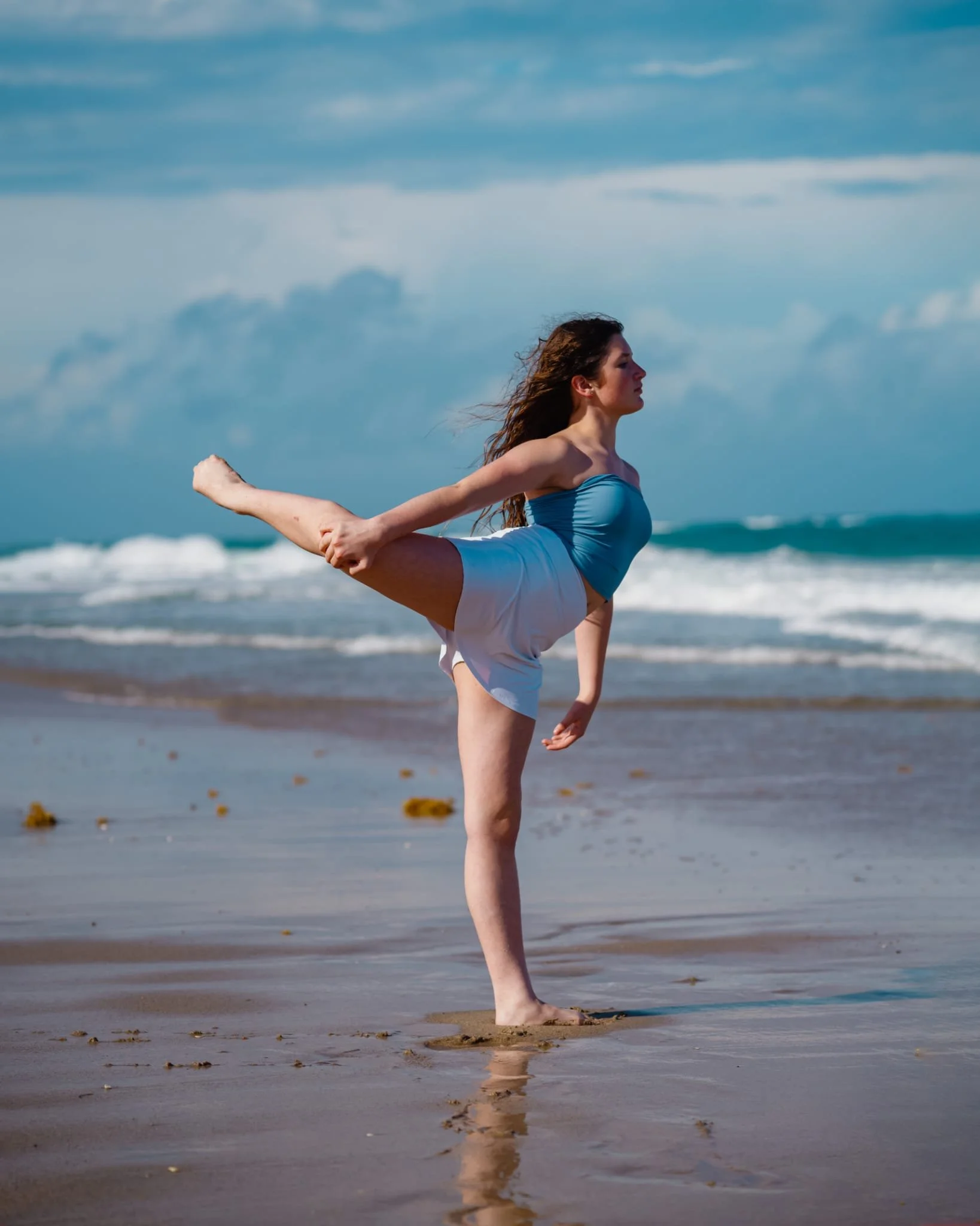 A young woman practicing dancing on the beach, standing on her left leg while holding her right leg behind her back with her right hand, overlooking the ocean with waves and cloudy sky.
