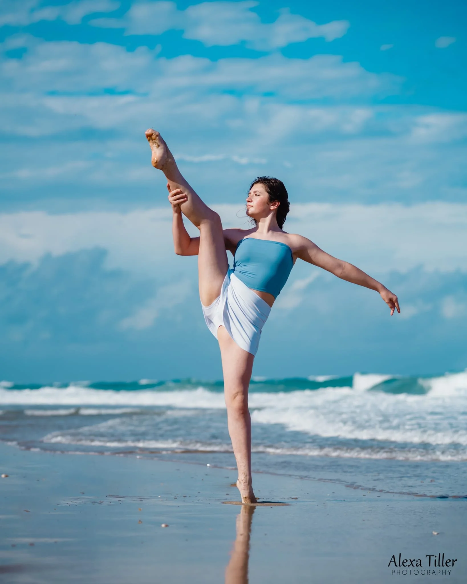 A young woman performing a dancing pose on the beach, standing on one leg with the other leg raised high and held by her hand, with ocean waves and blue sky in the background.