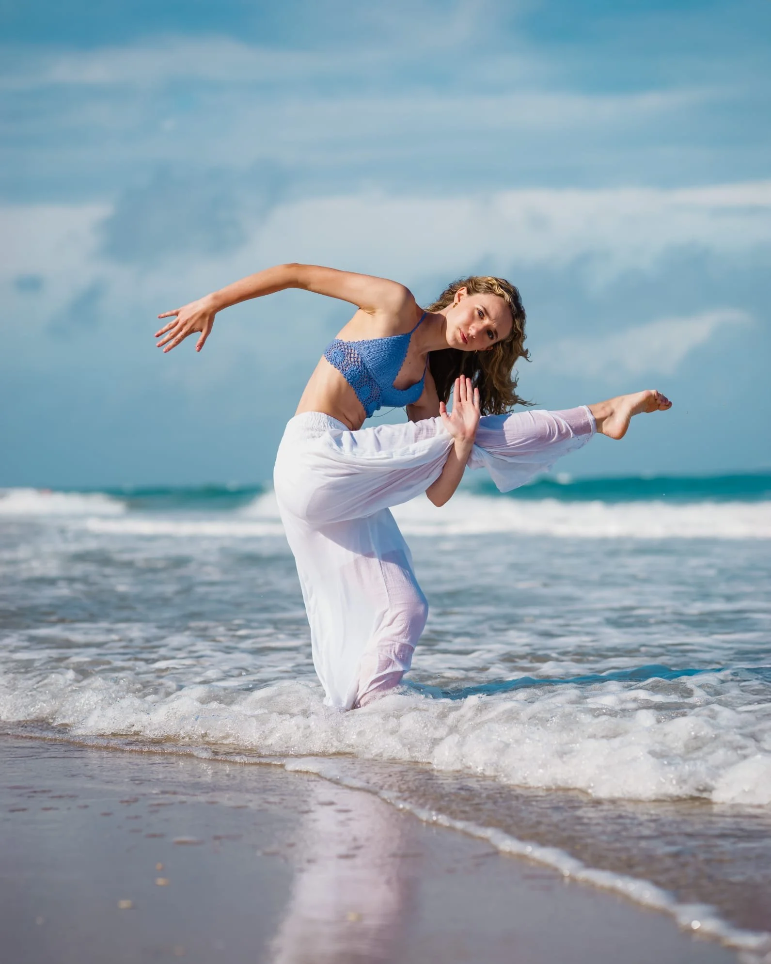 A young woman doing a dancing pose at the beach, standing in the shallow water with her leg lifted and hands together in prayer position.