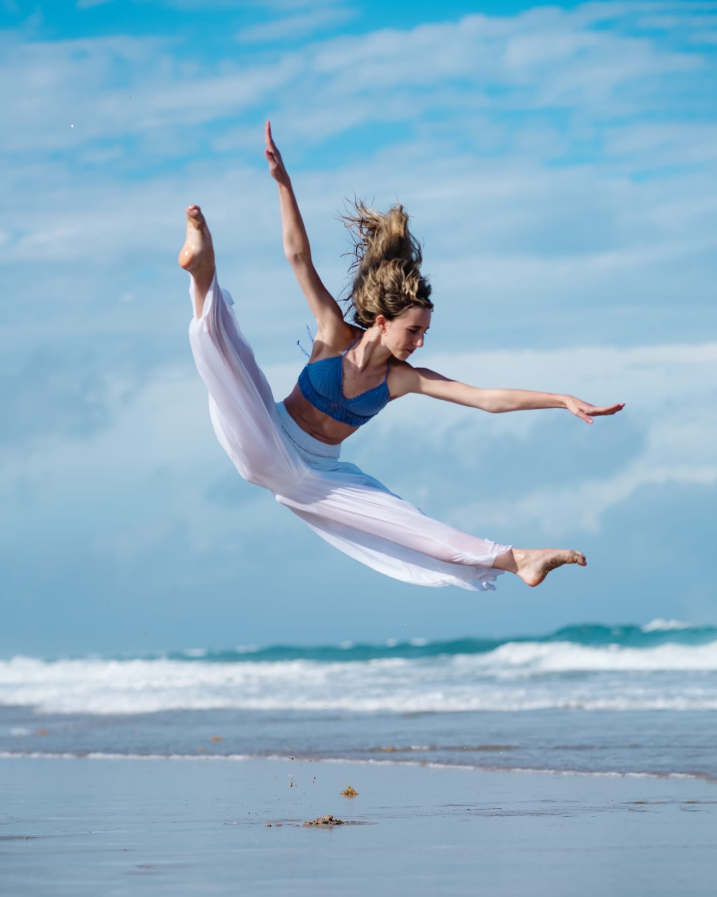 A young woman performing a dance jump on the beach with waves and blue sky in the background.