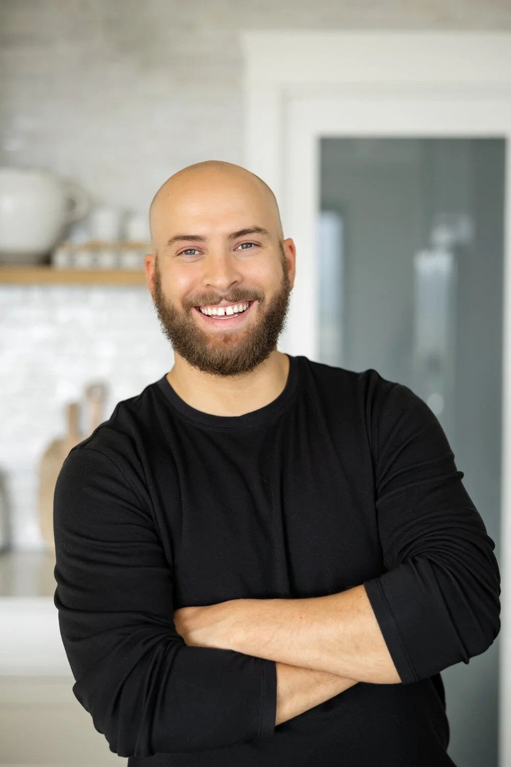 A smiling man with a beard and bald head, wearing a black long-sleeve shirt, standing with arms crossed in a modern kitchen with white cabinets and shelves.