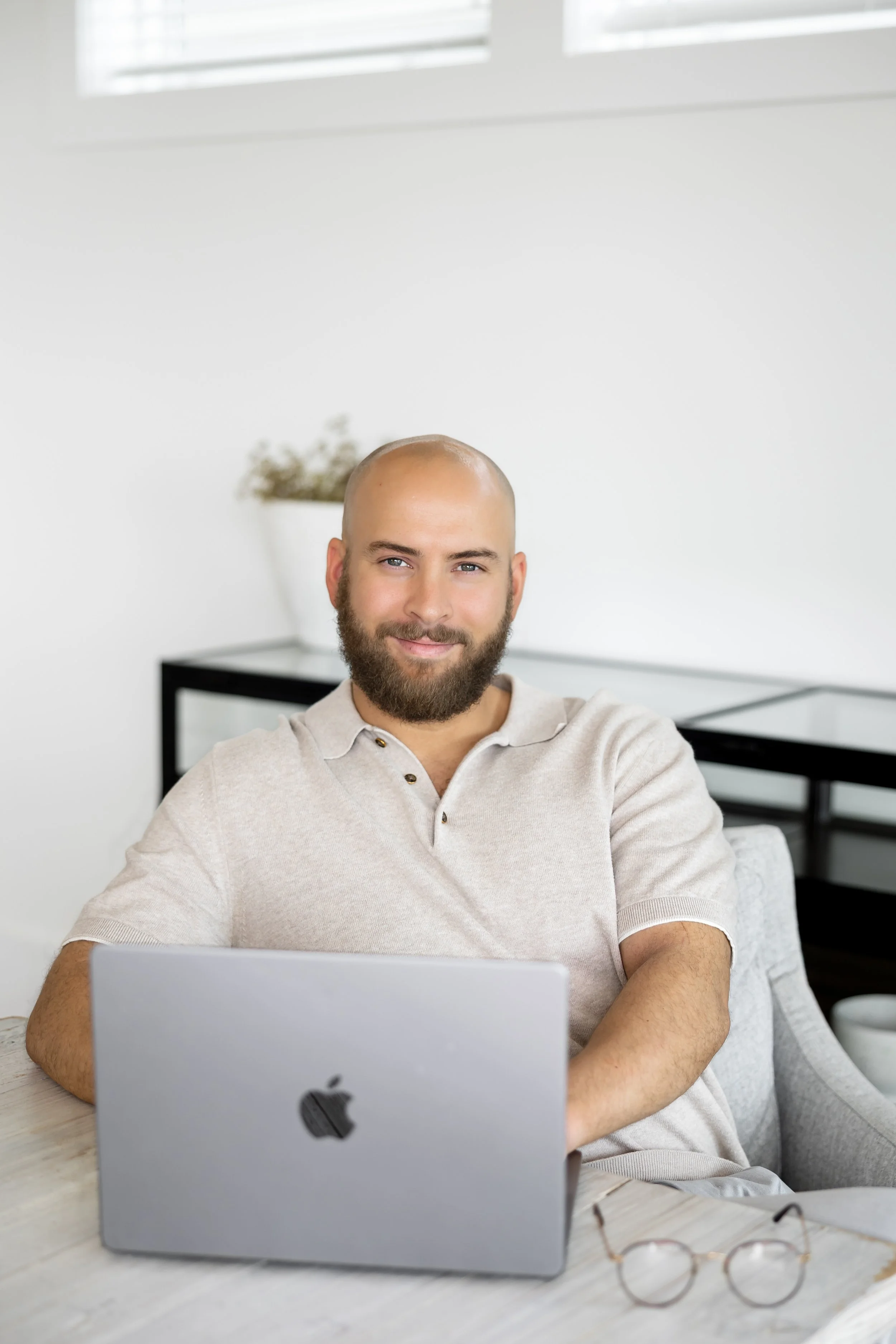 Man with a beard sitting at a desk with a silver Apple MacBook, smiling at the camera, in a bright, minimalist room.
