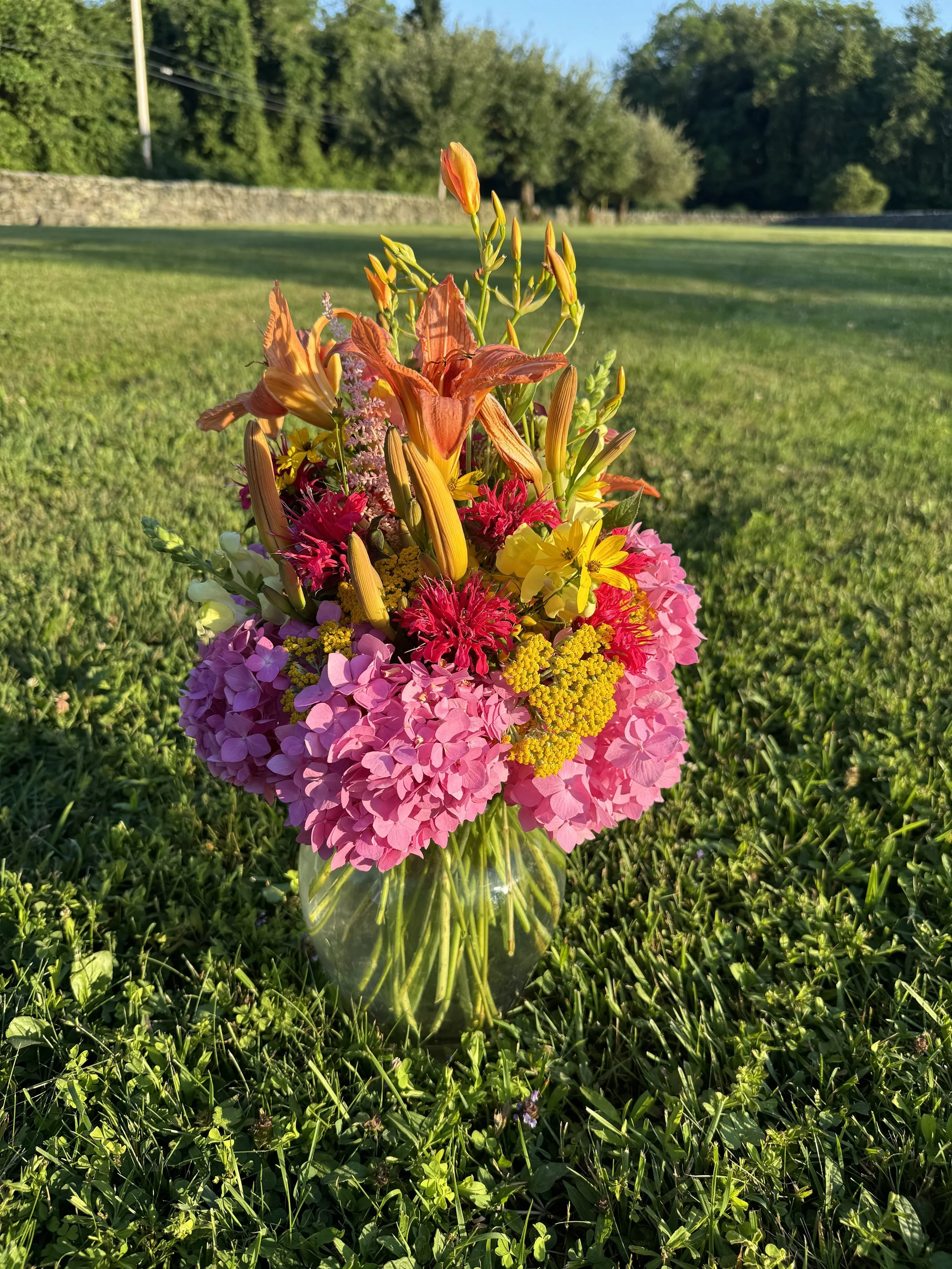 Colorful bouquet of pink, yellow, red, and orange flowers in a clear glass vase sitting on green grass with a background of trees and a stone wall.