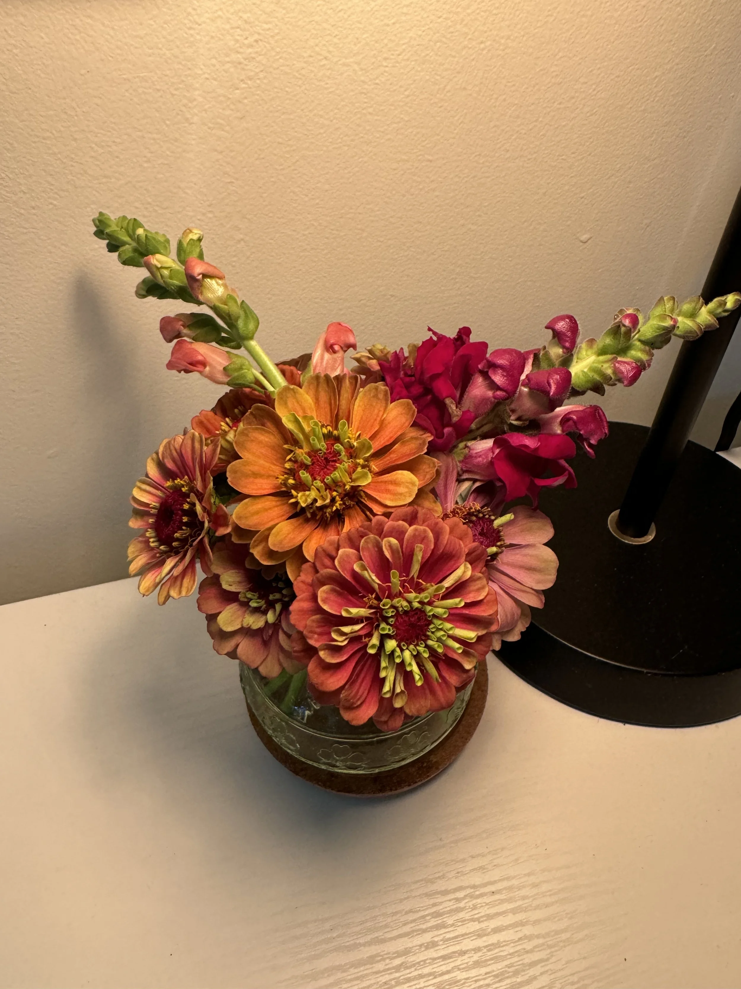A bouquet of colorful flowers in a glass vase on a white surface next to a black desk lamp.