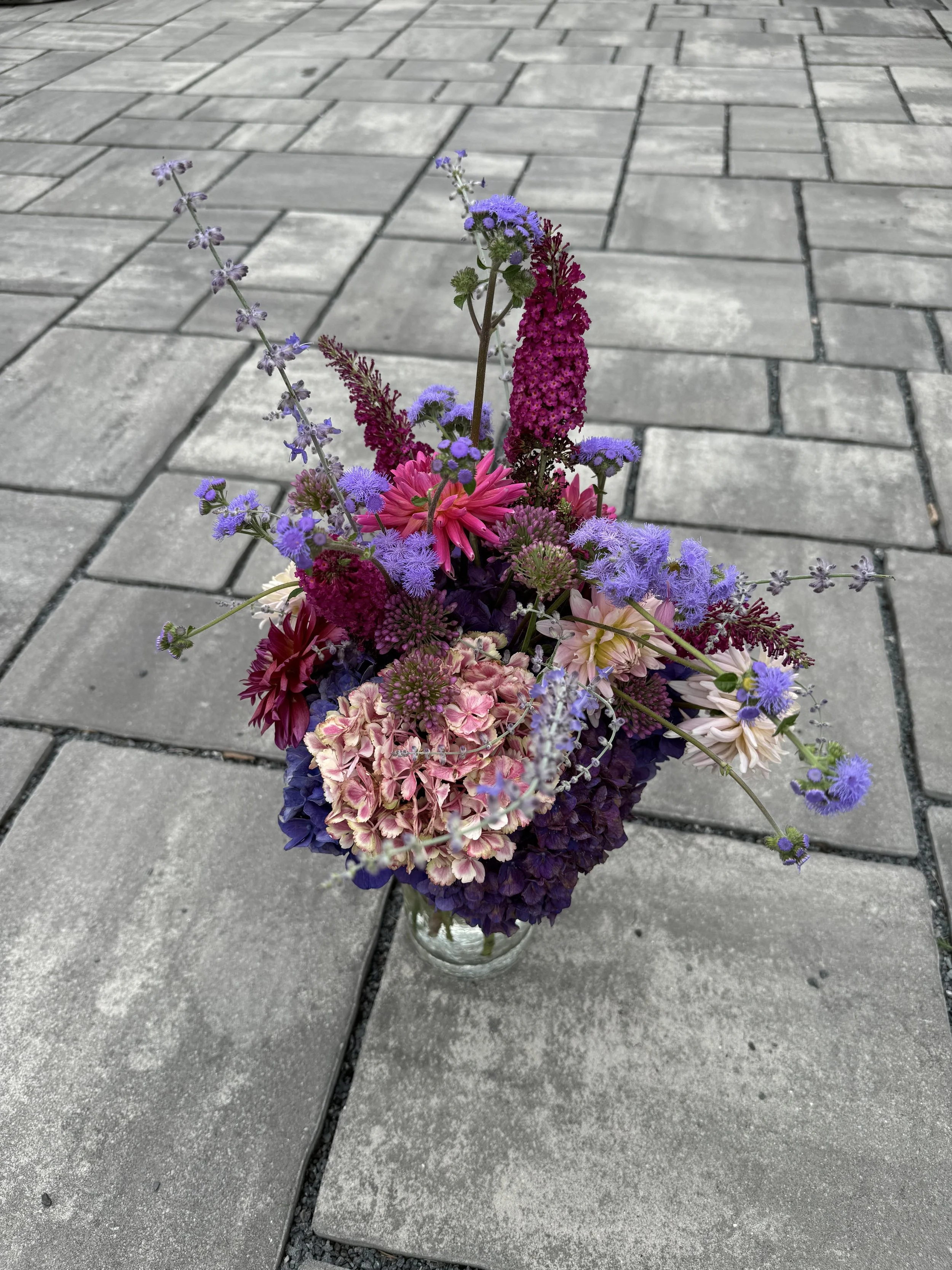 A colorful bouquet of flowers in a glass vase on a paved sidewalk with gray bricks.