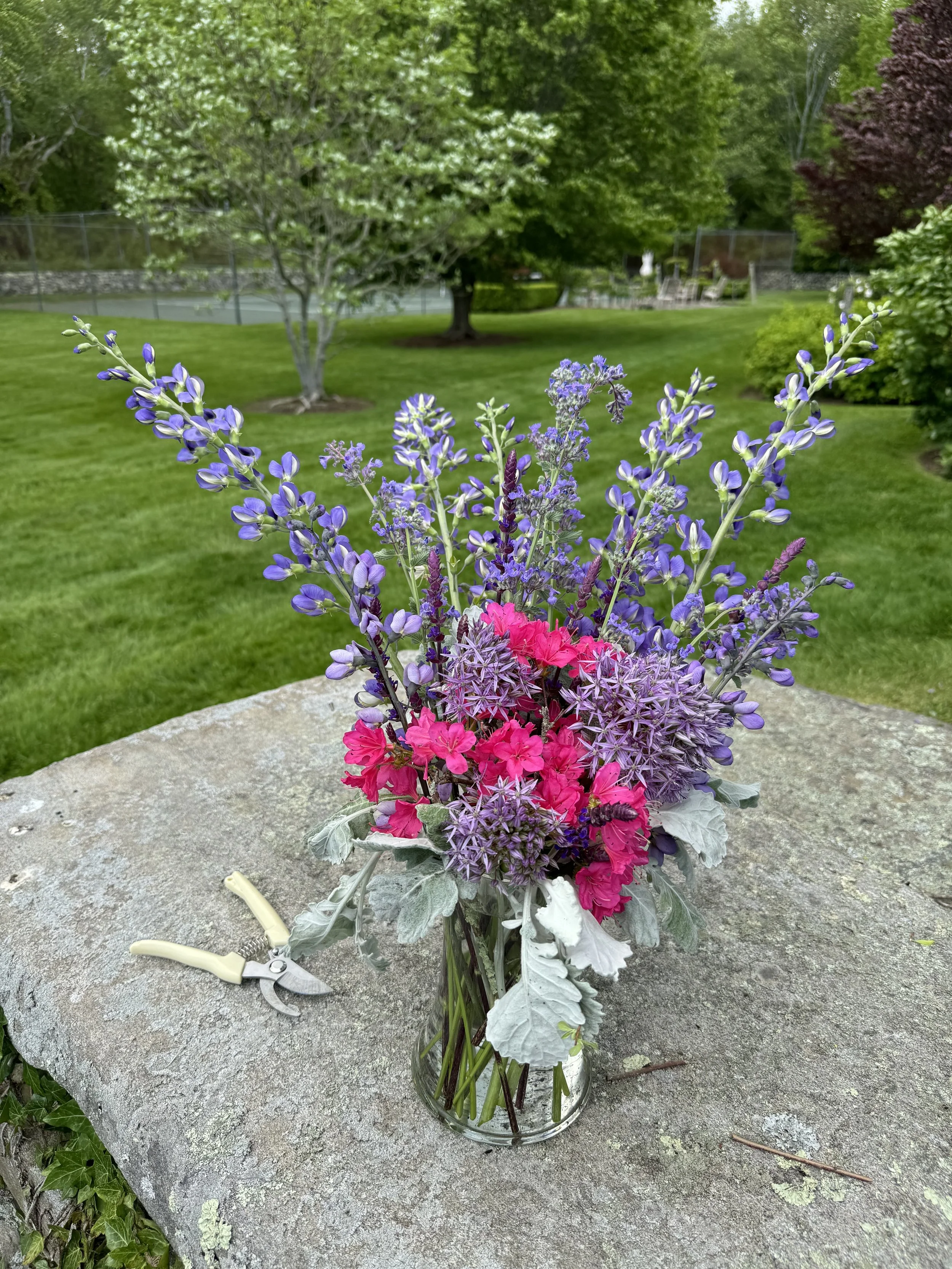 A colorful bouquet of purple, pink, and lavender flowers in a glass vase sits on a stone surface outdoors in a green, well-maintained yard with trees and a fence in the background.