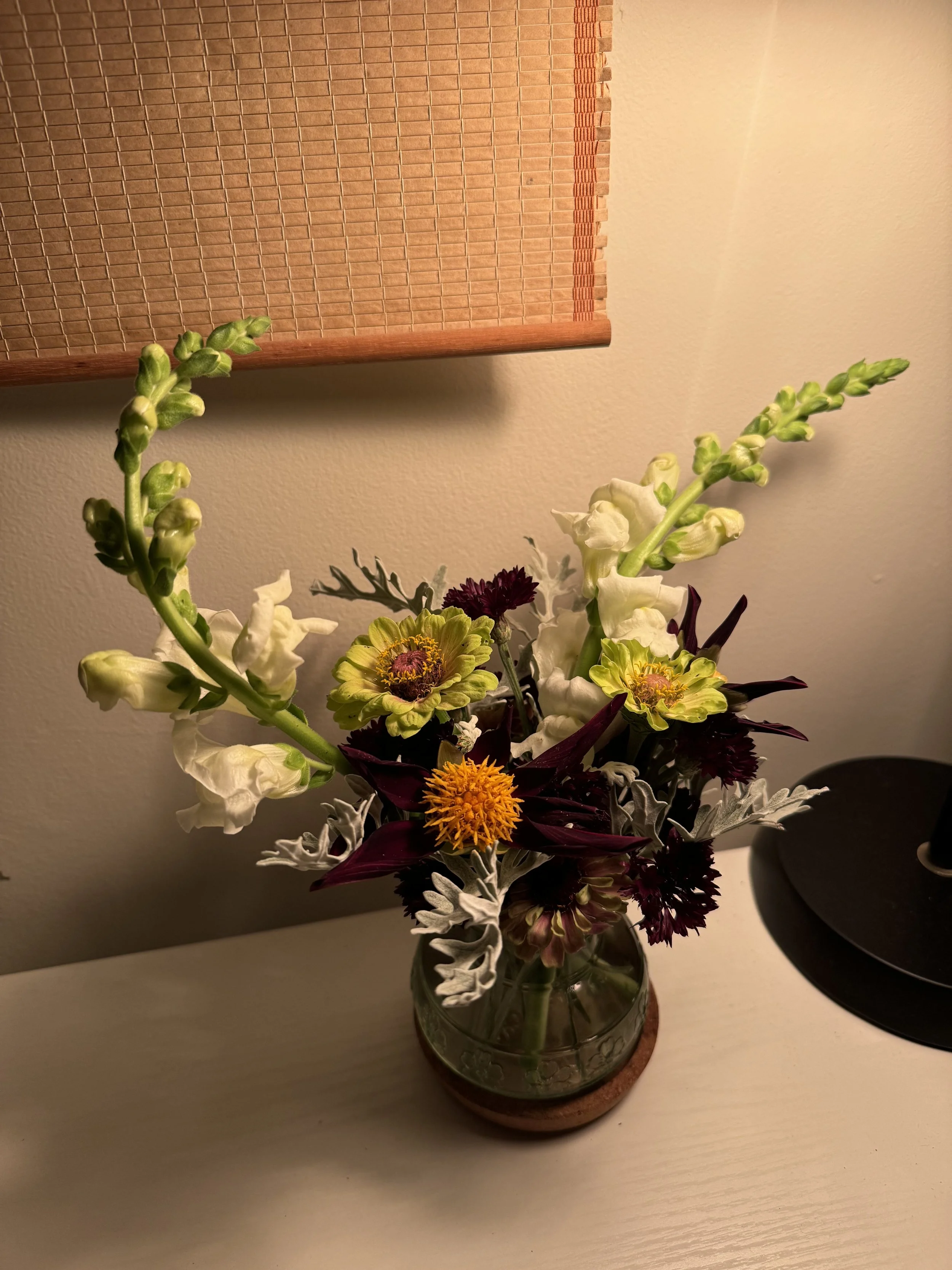 A vase of mixed flowers including white gladiolus, yellow and burgundy zinnias, orange pom-pom chrysanthemum, and dusty miller leaves on a white surface next to a black lamp against a beige wall.