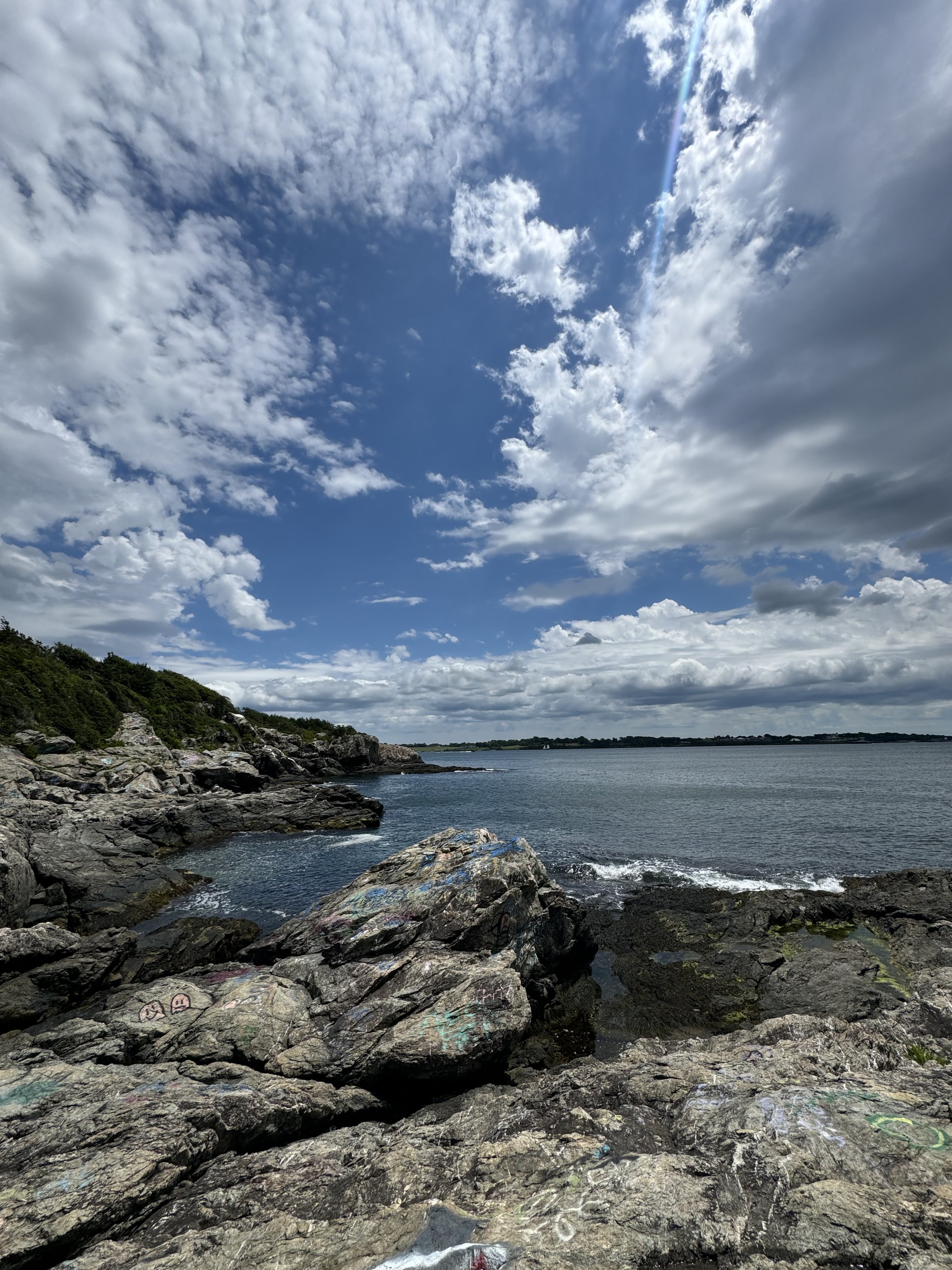 Rocky shoreline with graffiti, calm water, and a partly cloudy sky with the sun shining.