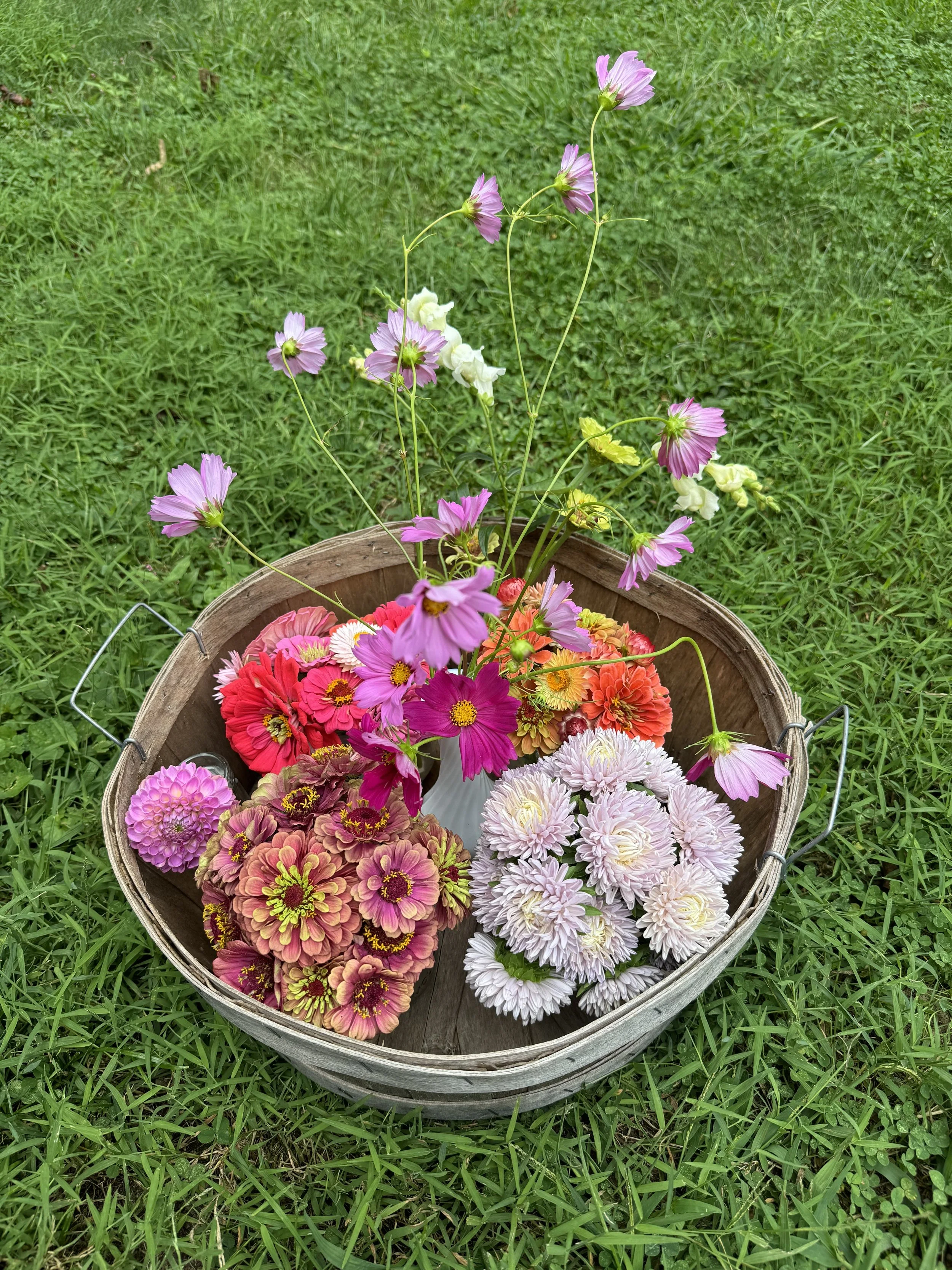 A wooden basket filled with various colorful flowers, including pink, white, and red blooms, sitting on green grass.