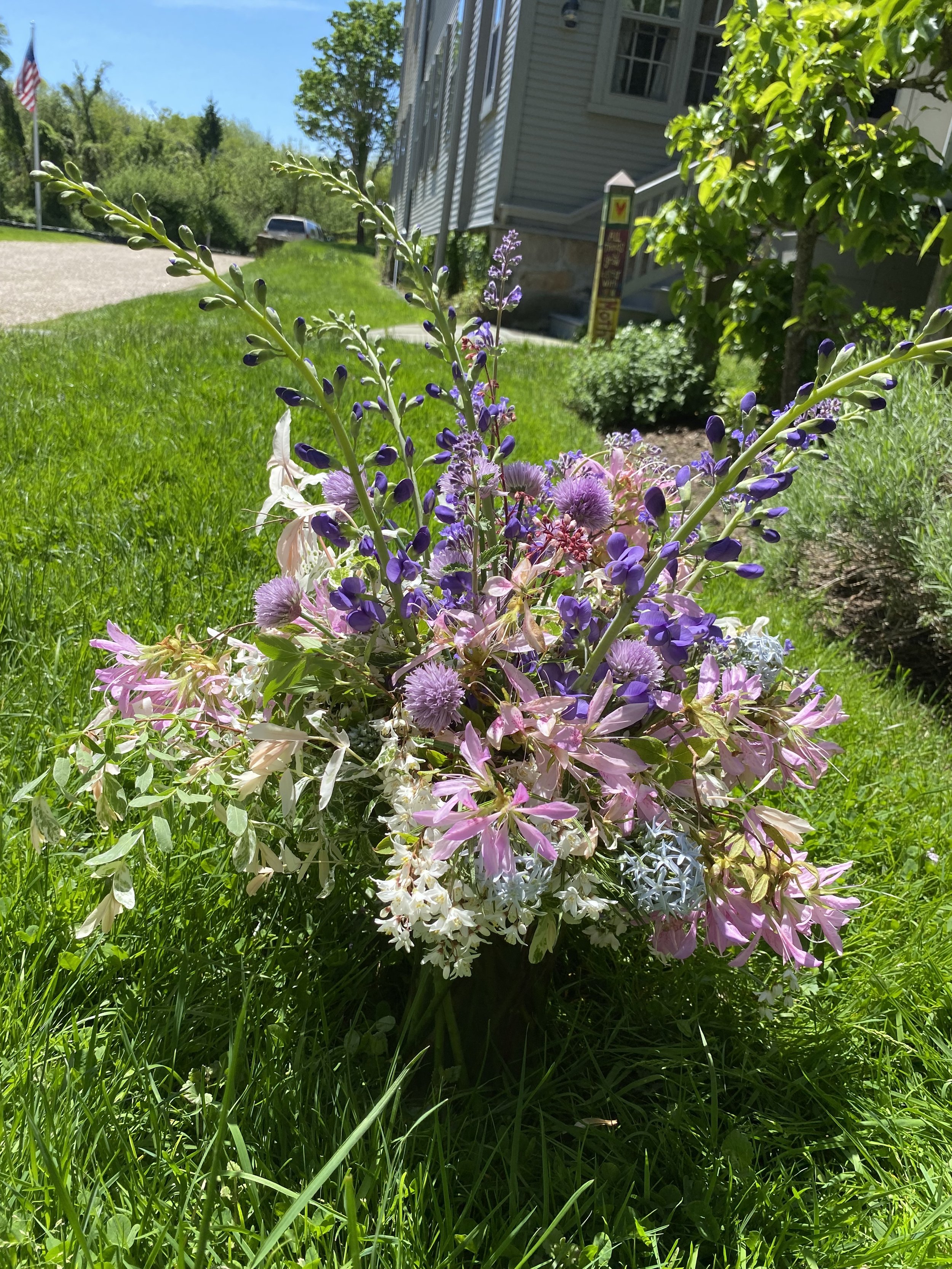 Brightly colored bouquet of mixed flowers, including purple, pink, and white blossoms, placed on green grass in front of a house on a sunny day.