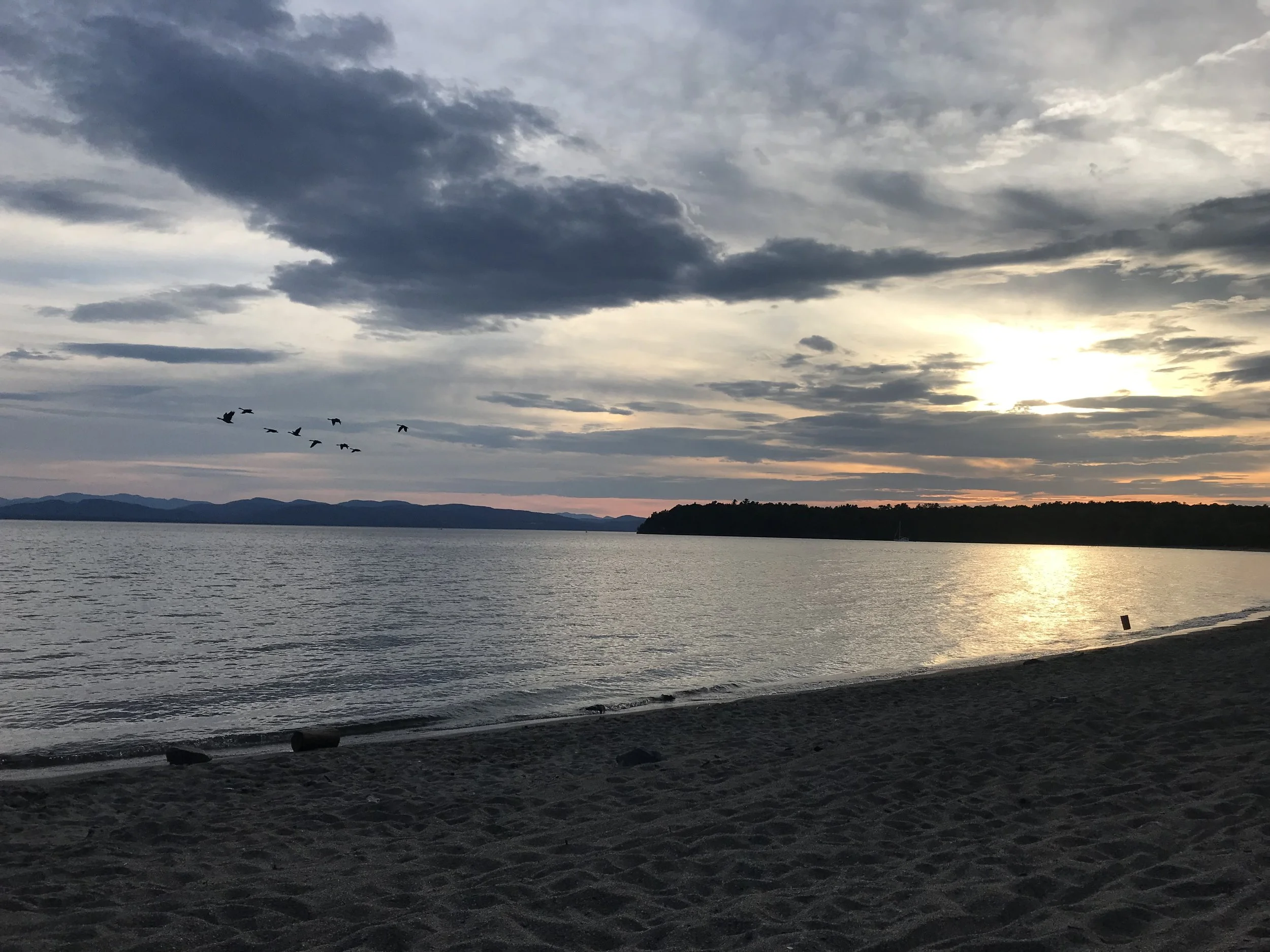 Sunset over a lake with a beach in the foreground, a group of flying birds, and a sailboat near the shoreline with mountains in the background.