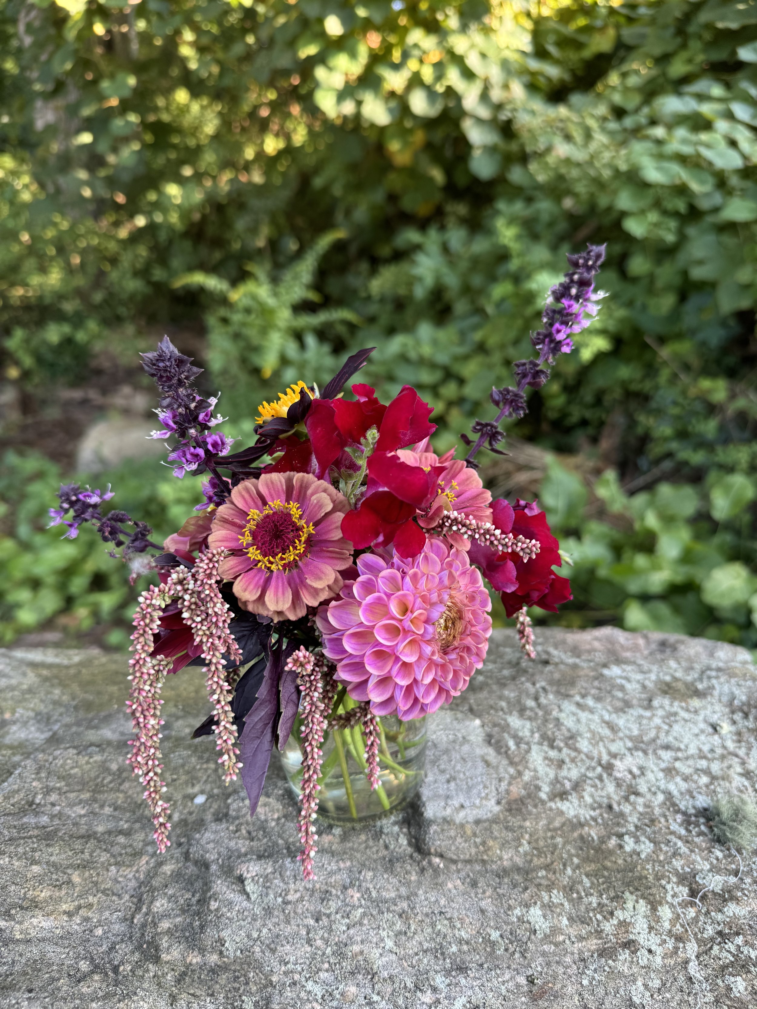 A colorful bouquet of various flowers in a glass jar, placed on a stone surface outdoors with greenery in the background.