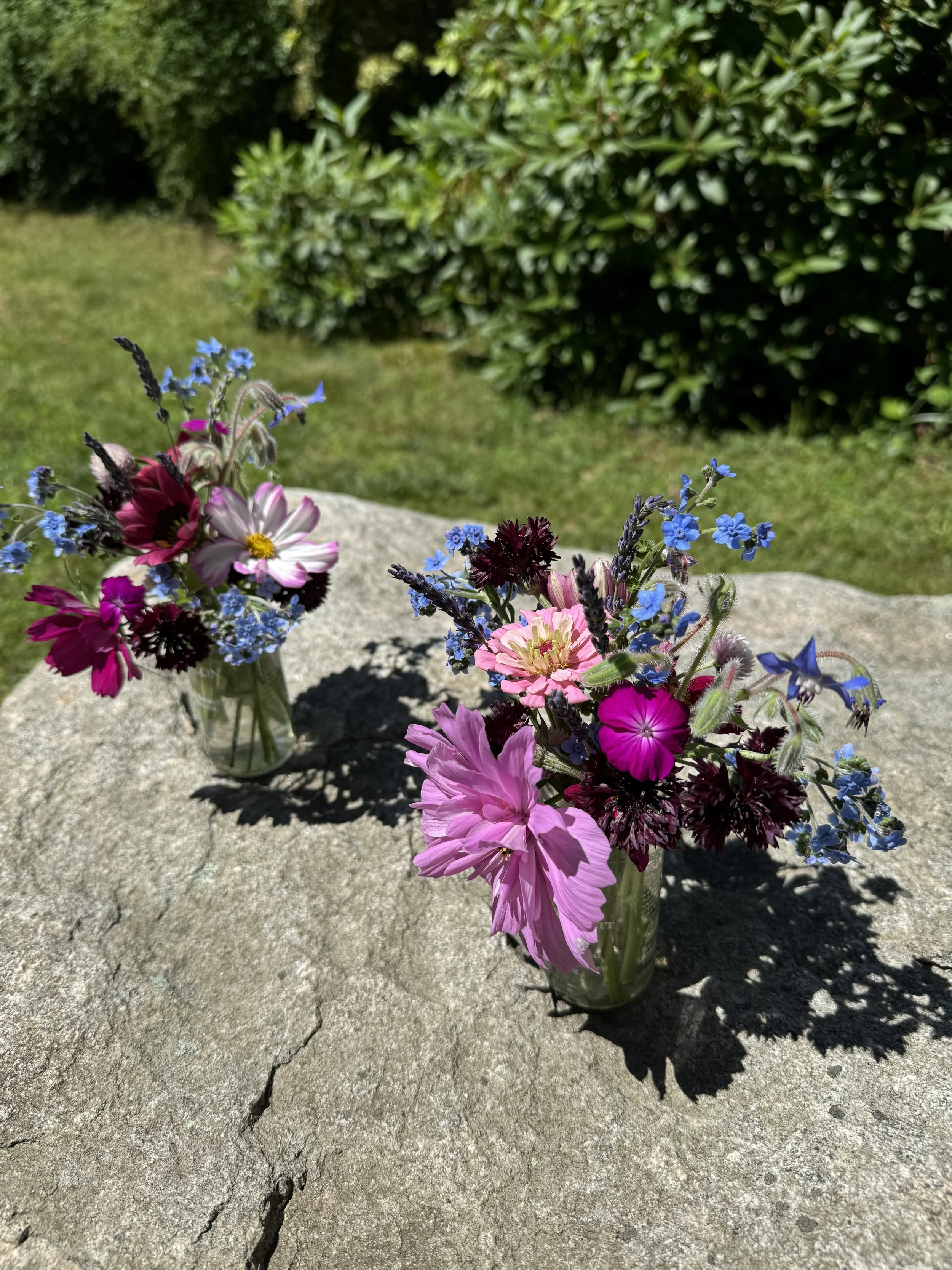 Two small glass jars filled with colorful wildflowers placed on a large stone outdoors.