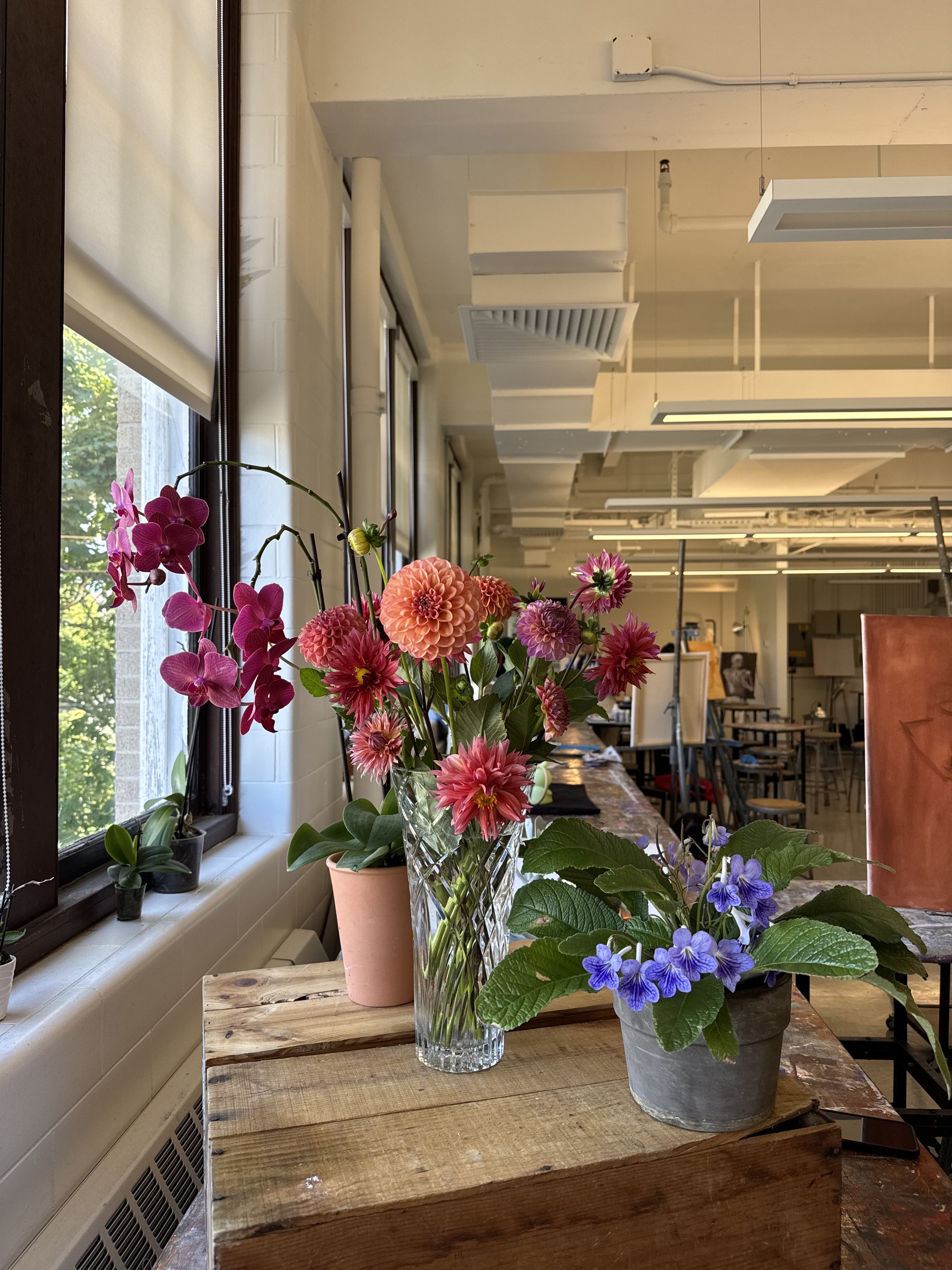 A wooden table with a vase of pink and red flowers and a potted purple flower, positioned near a window in a well-lit room with a white interior.