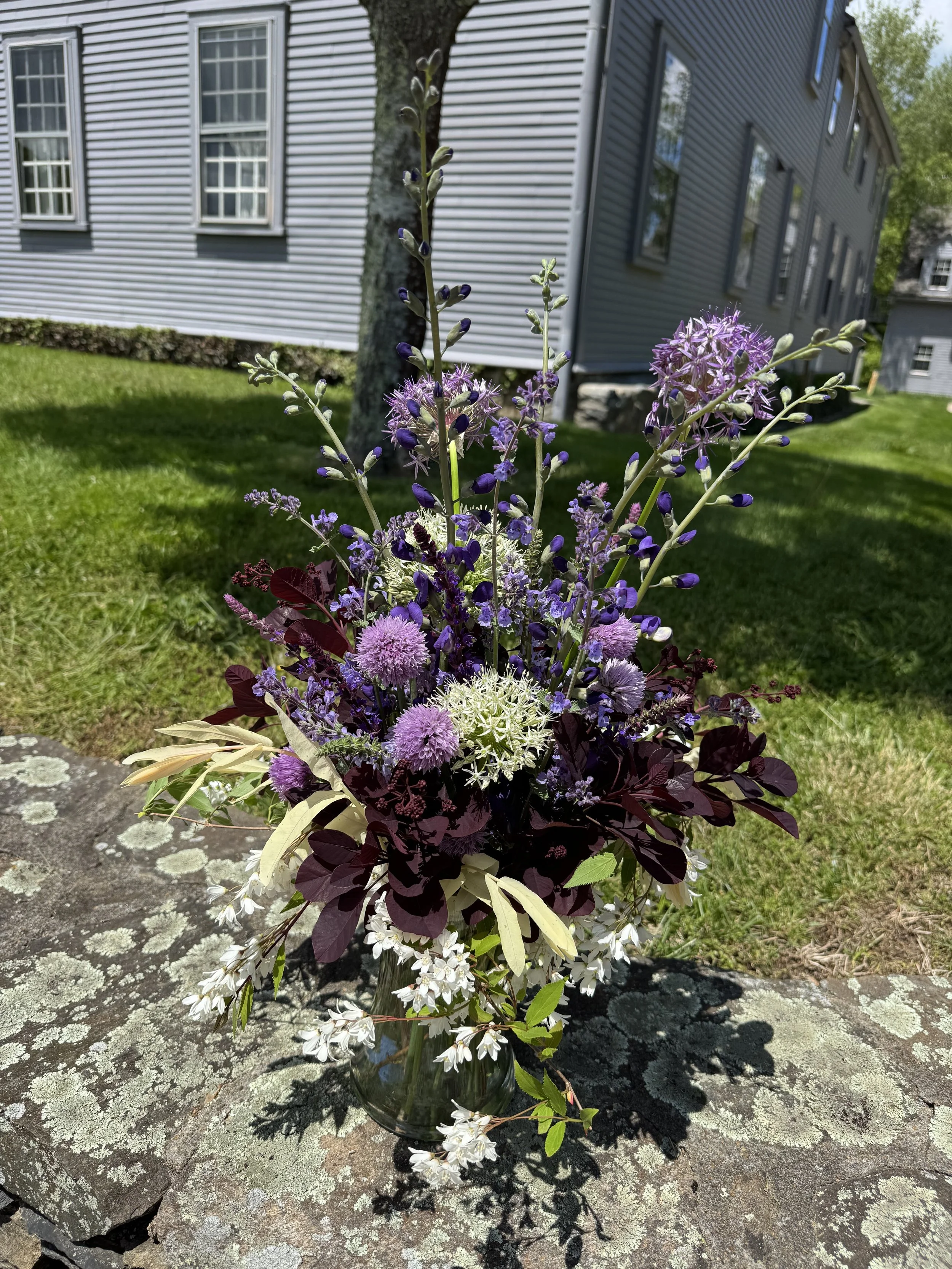 A bouquet of purple, white, and dark red flowers in a glass vase on a stone surface outdoors, with a house and green lawn in the background.