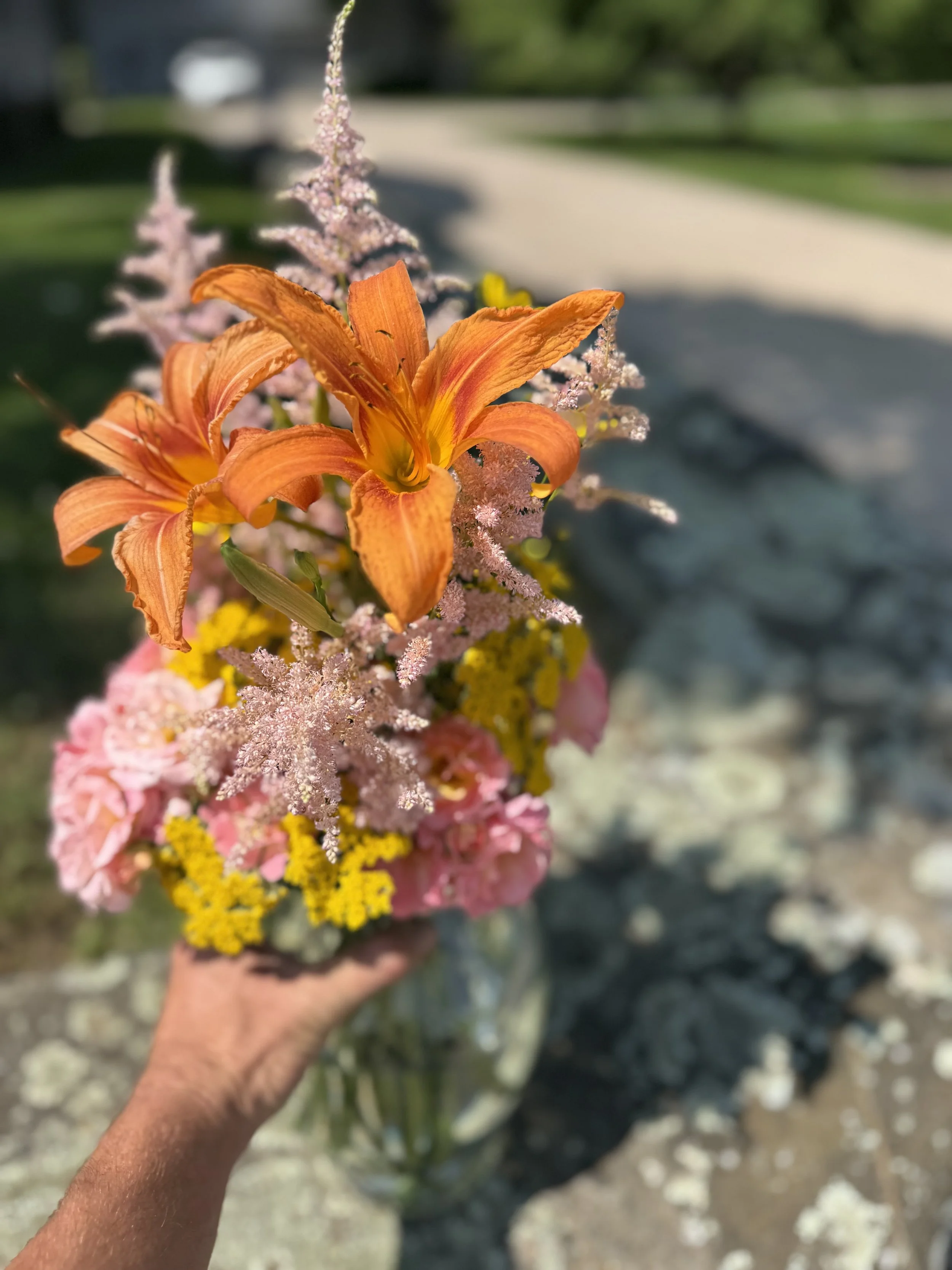 A hand holding a glass jar filled with colorful flowers, including orange lilies, pink hydrangeas, yellow filler flowers, and pink astilbes, outdoors with blurred greenery and a paved path in the background.