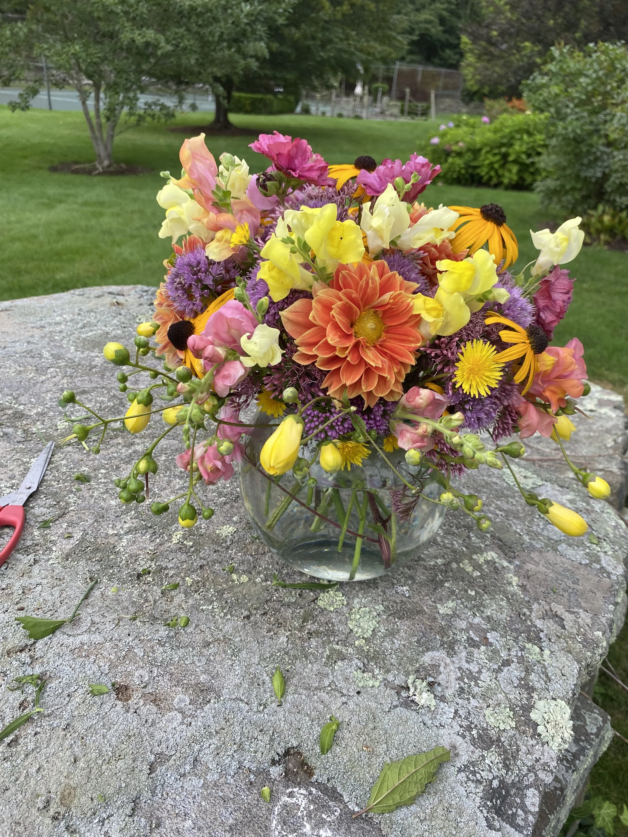 Colorful flower bouquet in a glass vase on a stone outdoor surface with a green yard and trees in the background.
