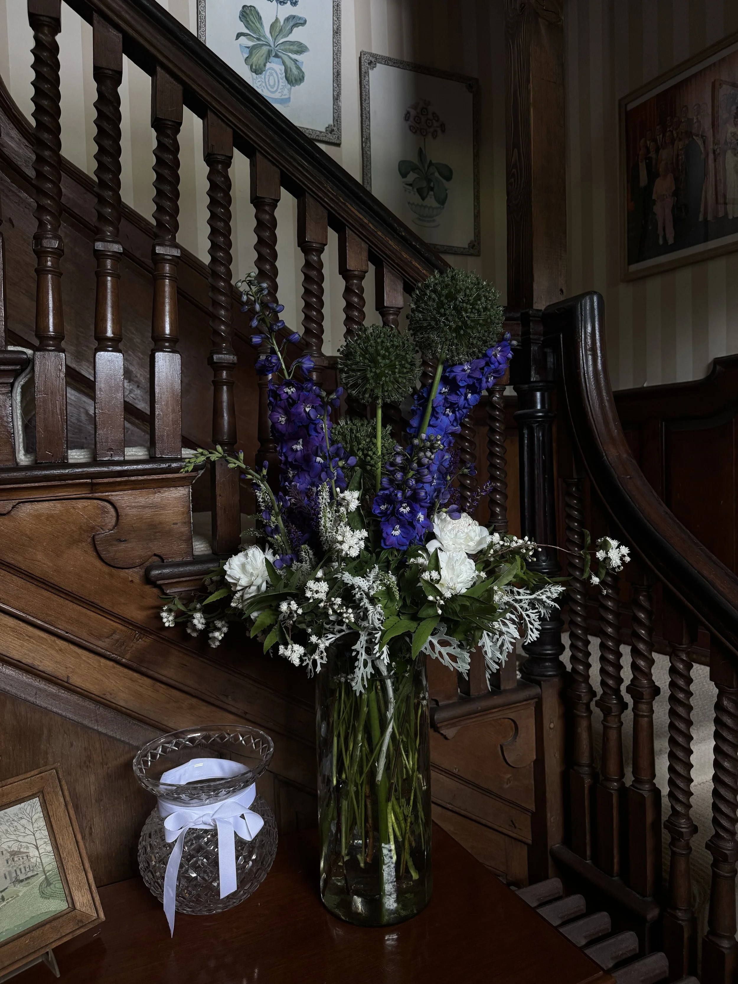 A large glass vase with white and purple flowers on a wooden table next to a small glass container with a white ribbon, in front of a wooden staircase with framed artwork on the wall.