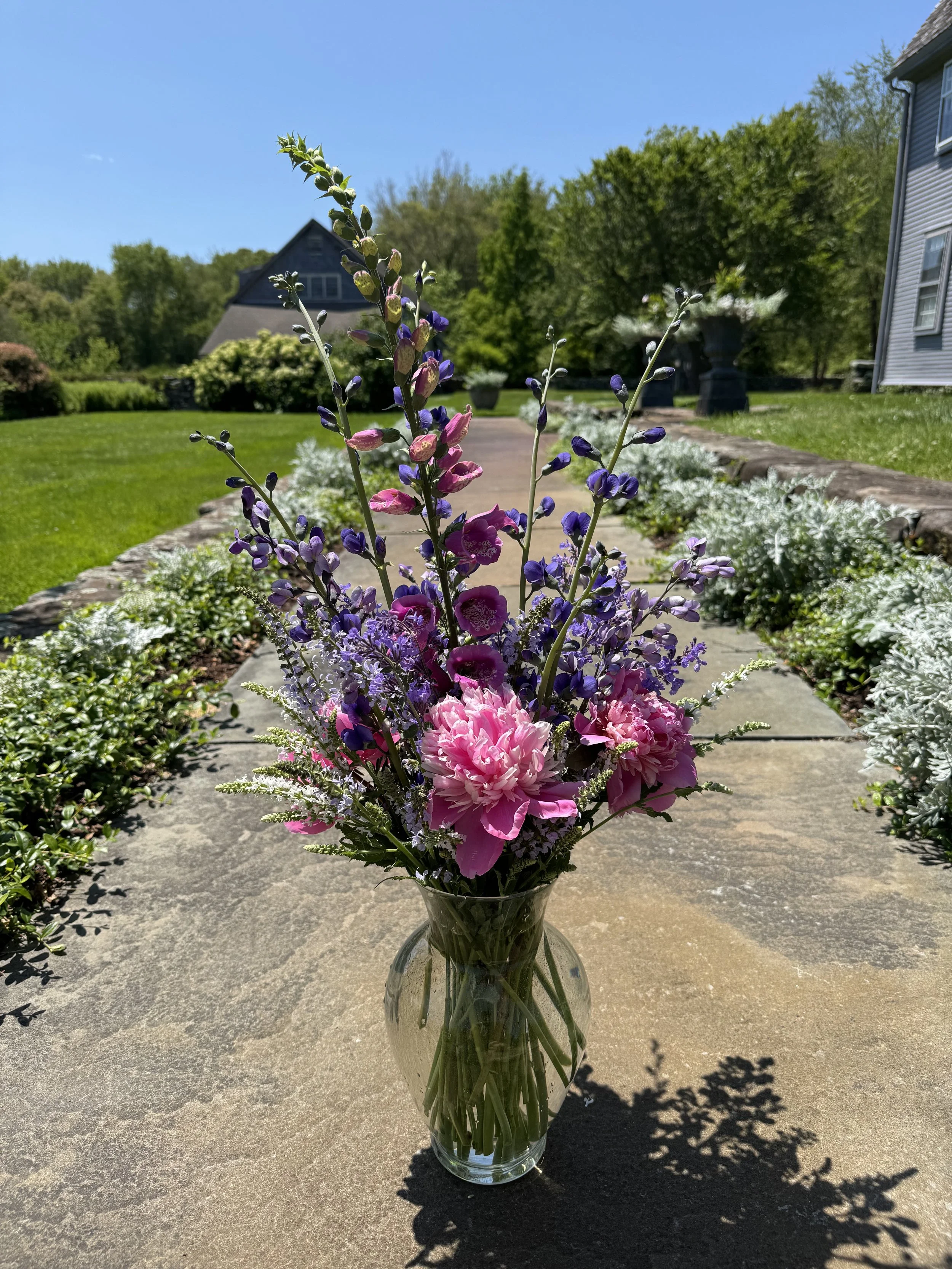 A glass vase with pink and purple flowers on a stone table outdoors, with a garden, trees, a house, and a blue sky in the background.