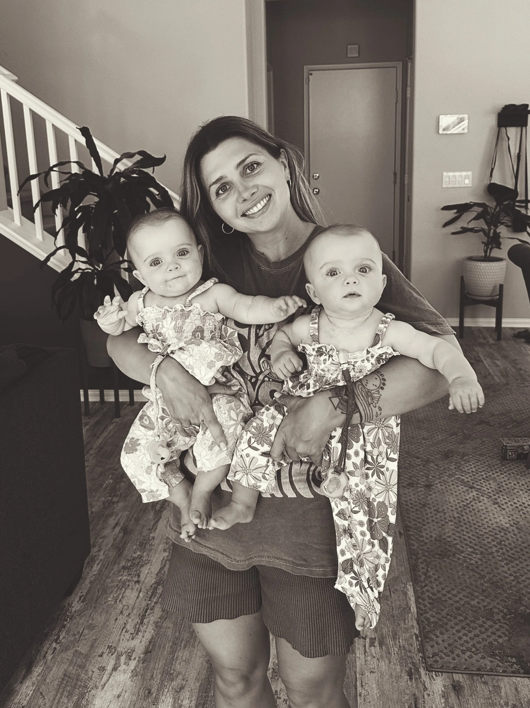 A woman smiling and holding two babies, standing indoors near a staircase and a potted plant.