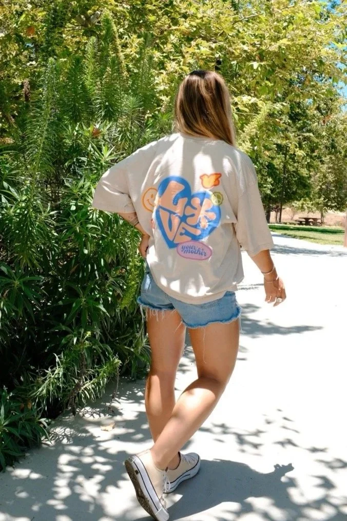 Person walking on a sunny outdoor path wearing a beige T-shirt with colorful graphics on the back, denim shorts, and white sneakers, surrounded by green trees and bushes.