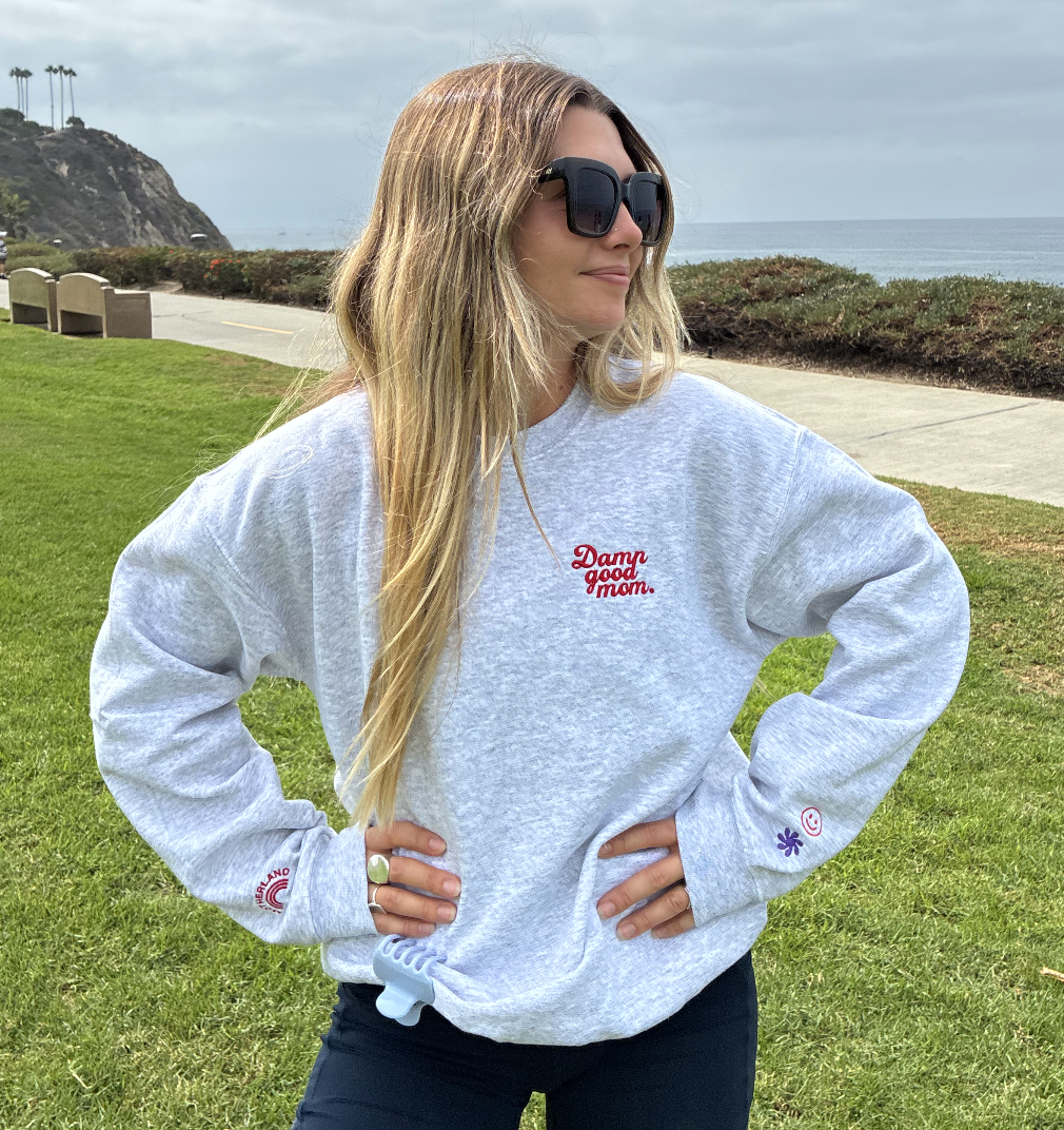 A woman with long blonde hair and sunglasses standing outdoors on a grassy area near the ocean, wearing a light gray sweatshirt with red and purple embroidered text and symbols, and dark pants.