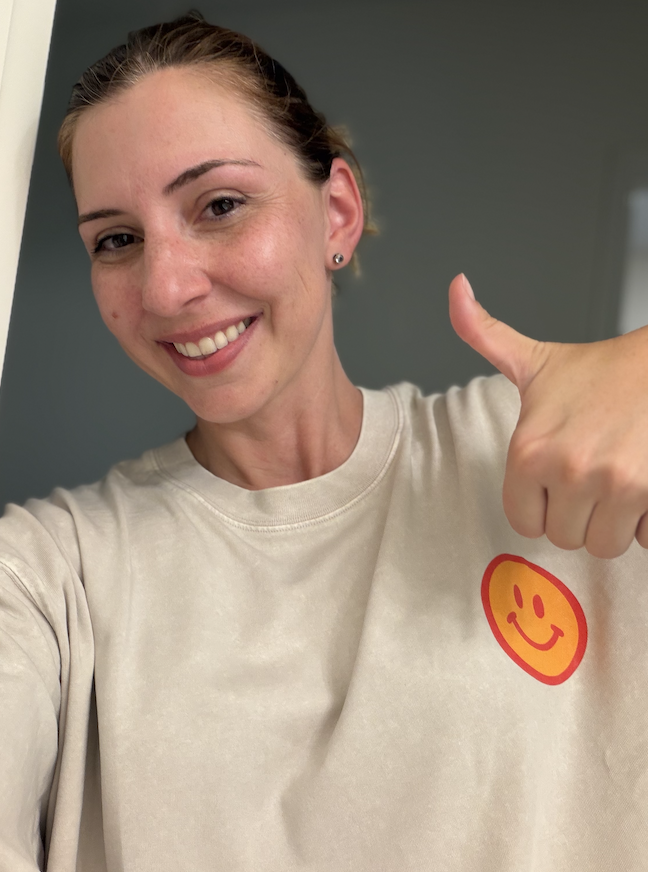 A woman smiling and giving a thumbs-up, wearing a beige T-shirt with a red and yellow smiley face logo.