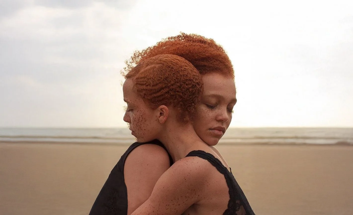 Two sisters with curly red hair hugging on a beach, with an overcast sky and grey sea in the background.