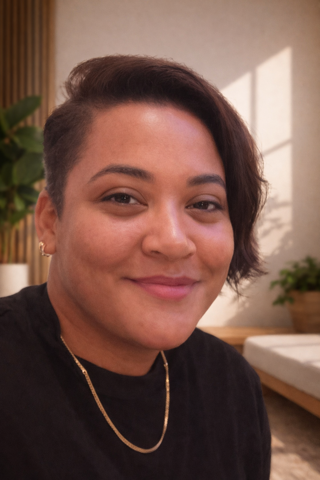 Tash, close-up photo of a smiling woman with short, dark hair, wearing a black top, gold chain necklace, and earrings, indoors with a plant and wooden elements in the background.