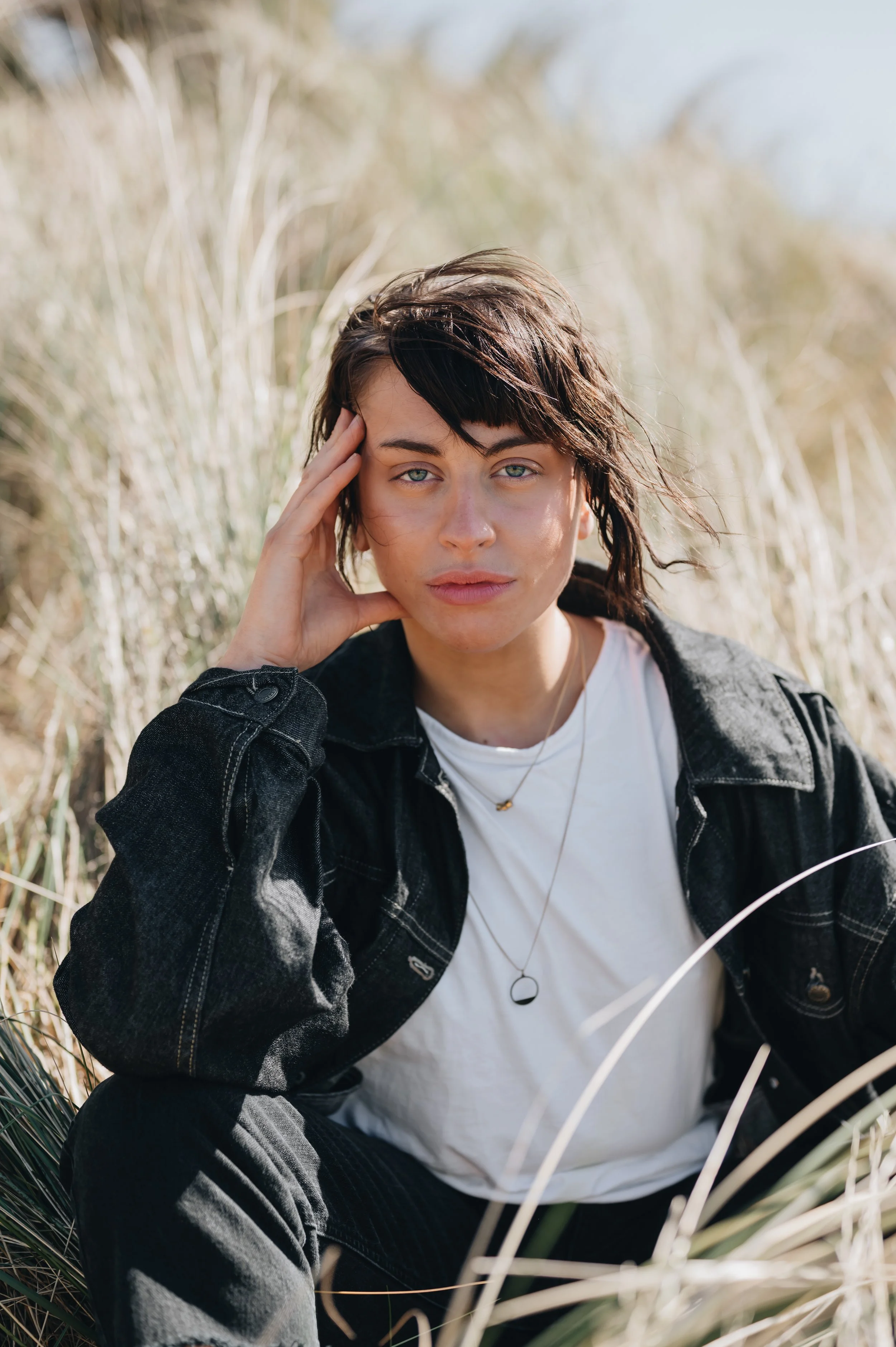 A young woman with short dark hair and light skin, sitting outdoors among tall dry grass, looking directly at the camera with a serious expression, wearing a black denim jacket and a white t-shirt, with her hand resting on the side of her head.