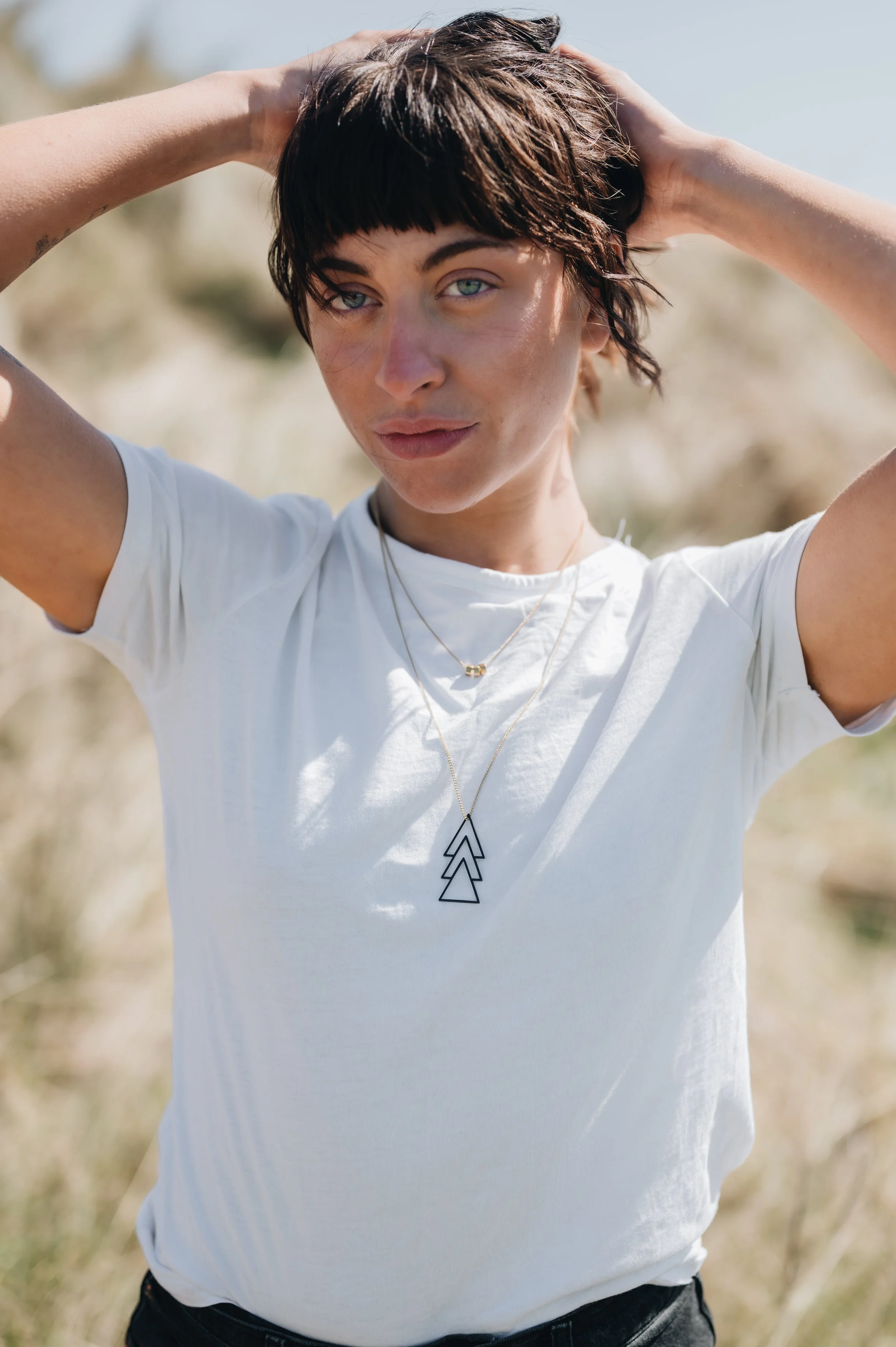 Woman with short dark hair and blue eyes, wearing a white t-shirt and layered necklaces, standing outdoors with her hands behind her head on a sunny day.
