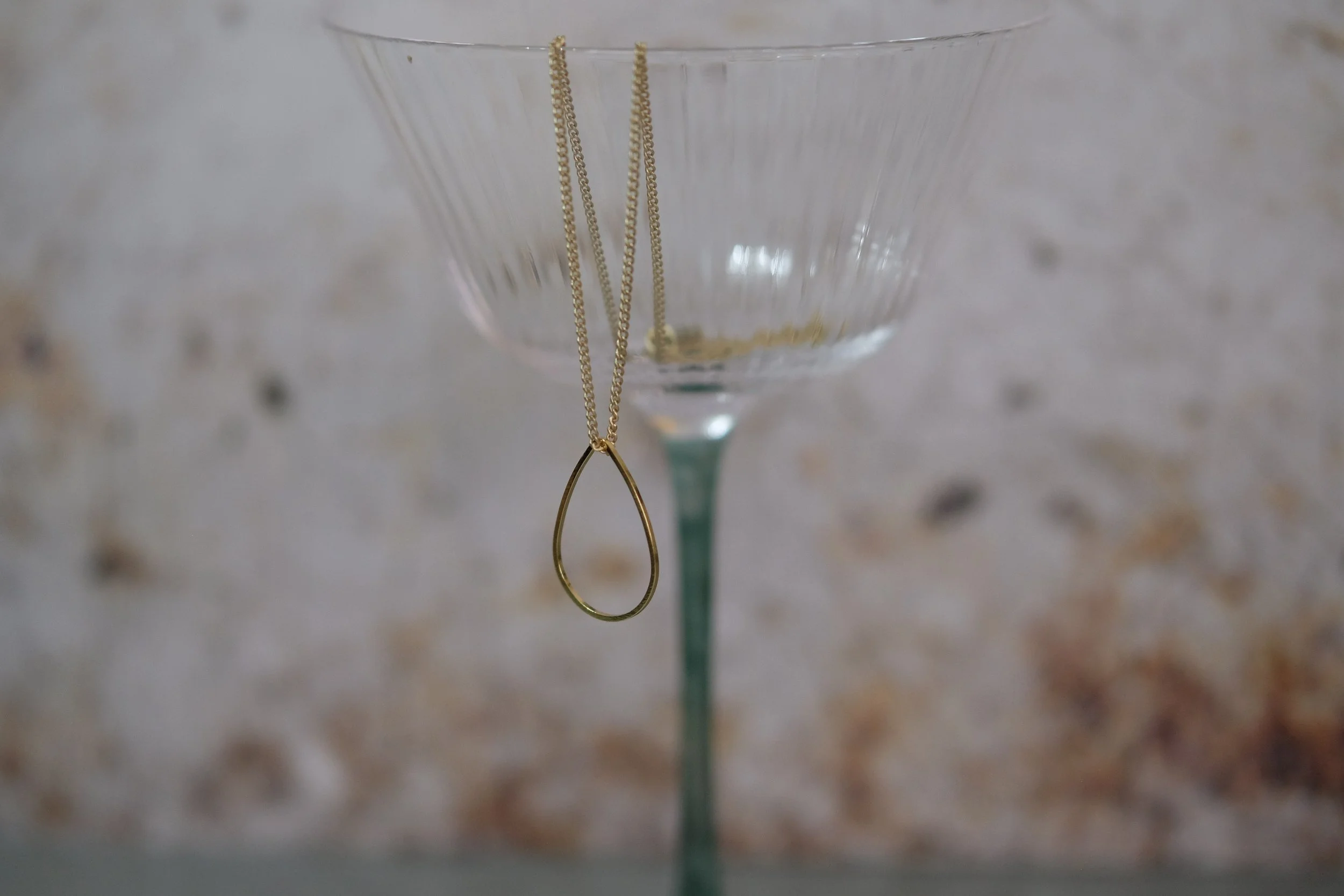 Gold necklace with a teardrop pendant hanging on the rim of a clear glass coupe with a green stem, against a textured background.