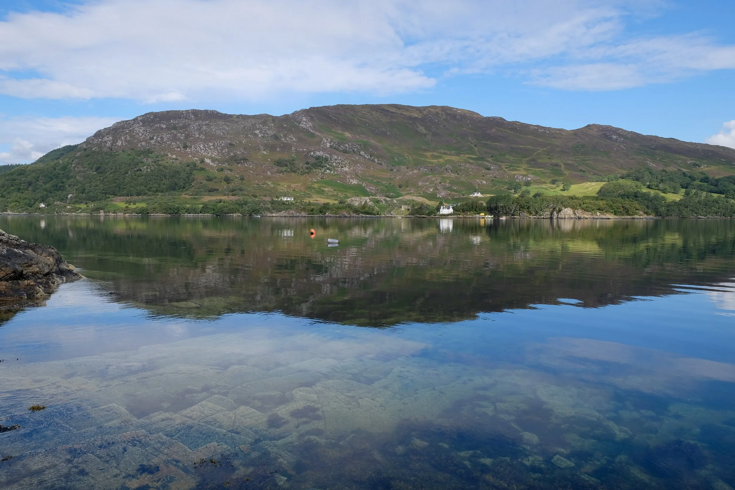 Scenic view of a calm lake with a mountain in the background, green hills, and a partly cloudy blue sky.