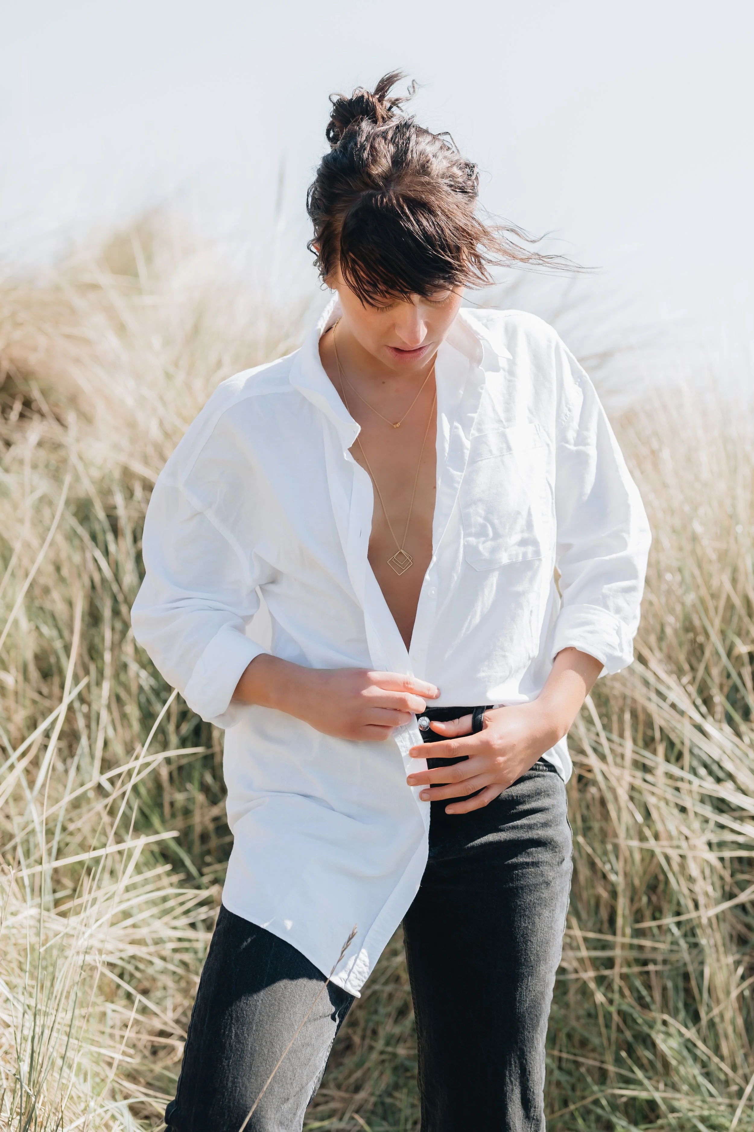 Woman with short dark hair wearing a white button-up shirt and dark pants standing in tall grass outdoors.