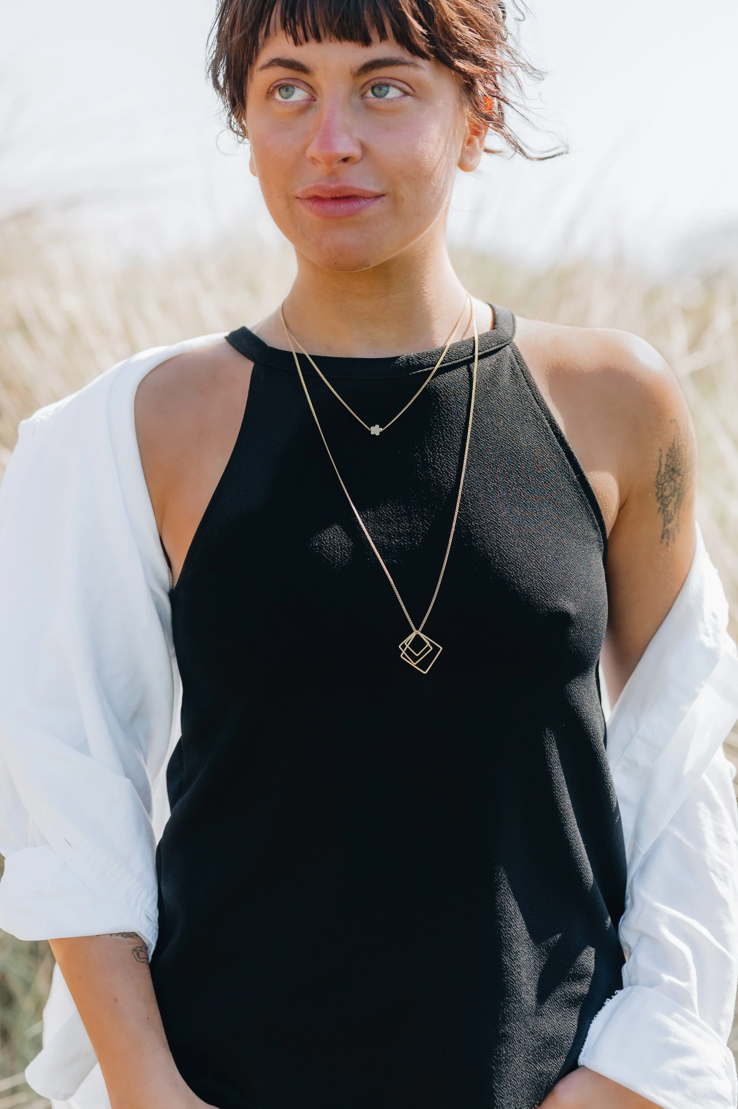 A woman with short brown hair wearing a black sleeveless top, layered necklaces, and a white shirt draped over her shoulders standing outdoors in a grassy field.