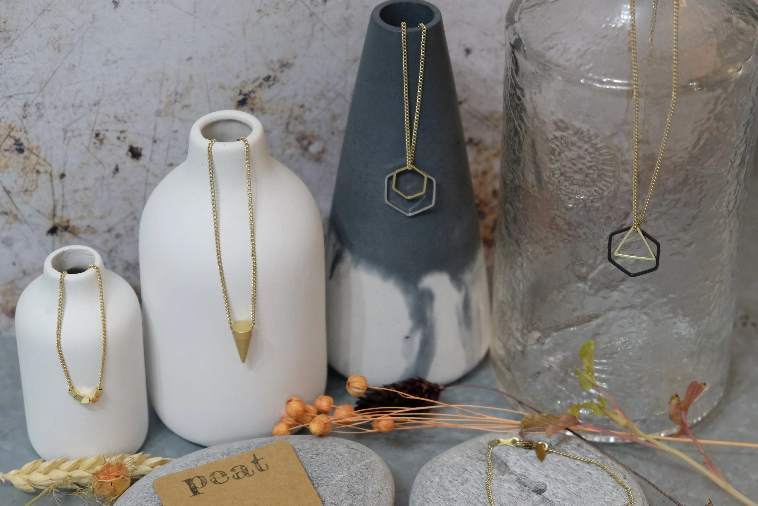 Display of five necklaces with geometric pendants on ceramic vases and a textured glass jar, surrounded by dried wheat and small orange berries, on a gray surface with a stone labeled "pearl" in the foreground.