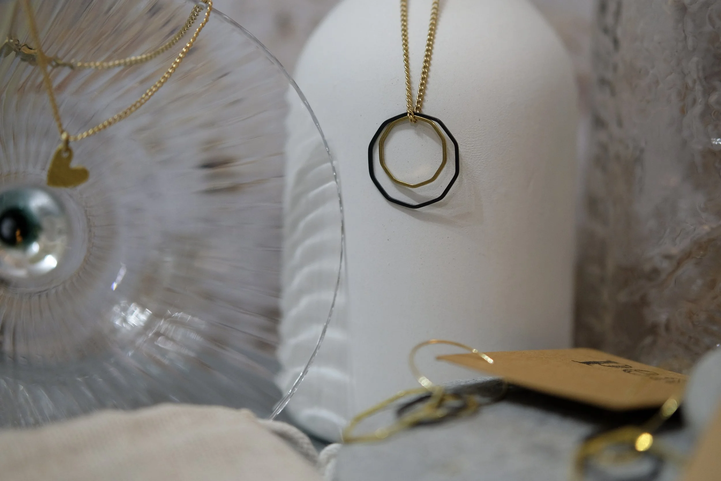 Several layered necklaces with different shaped pendants hanging against a white surface, next to a textured stone wall with a cardboard tag nearby.