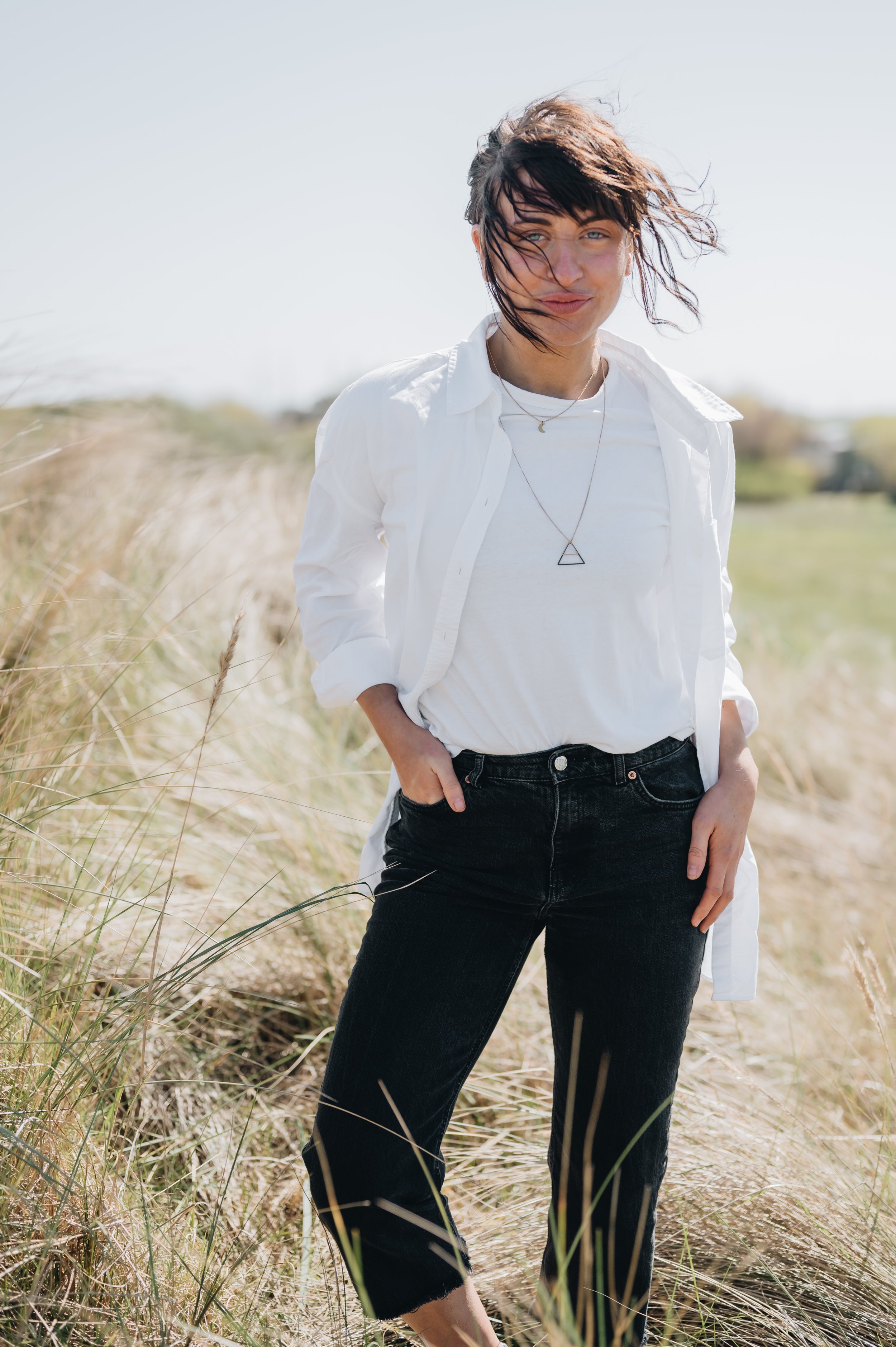 A young woman stands in a grassy field on a sunny day, wearing a white shirt, black jeans, and layered necklaces.