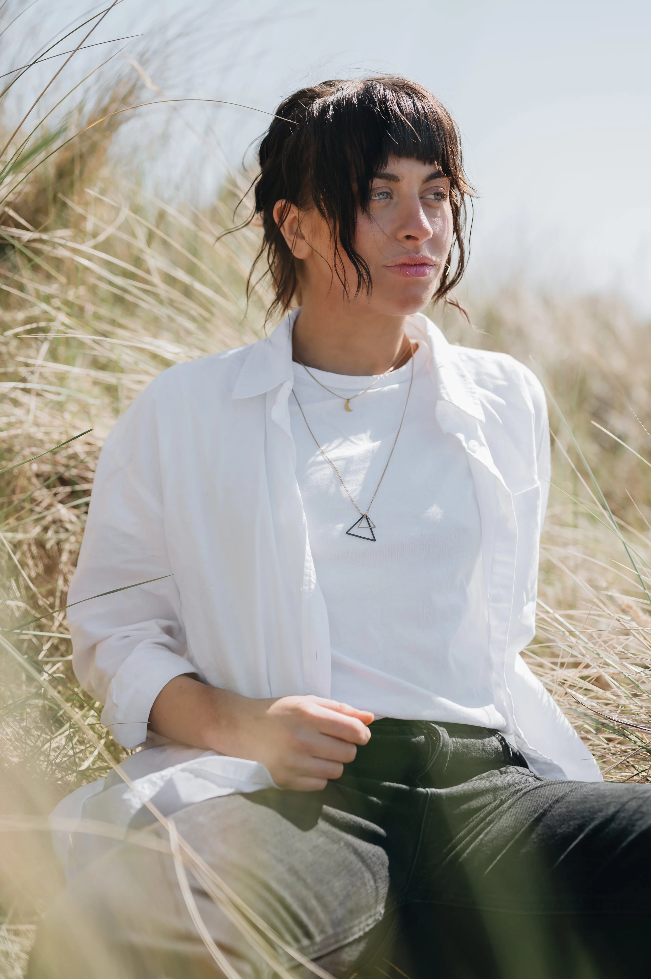 A woman with dark, wavy, short hair, sitting among tall, dry grass on a sunny day. She is wearing a white button-up shirt over a white t-shirt and black jeans. She has on layered necklaces, including one with a triangle pendant, and gazes thoughtfully into the distance.