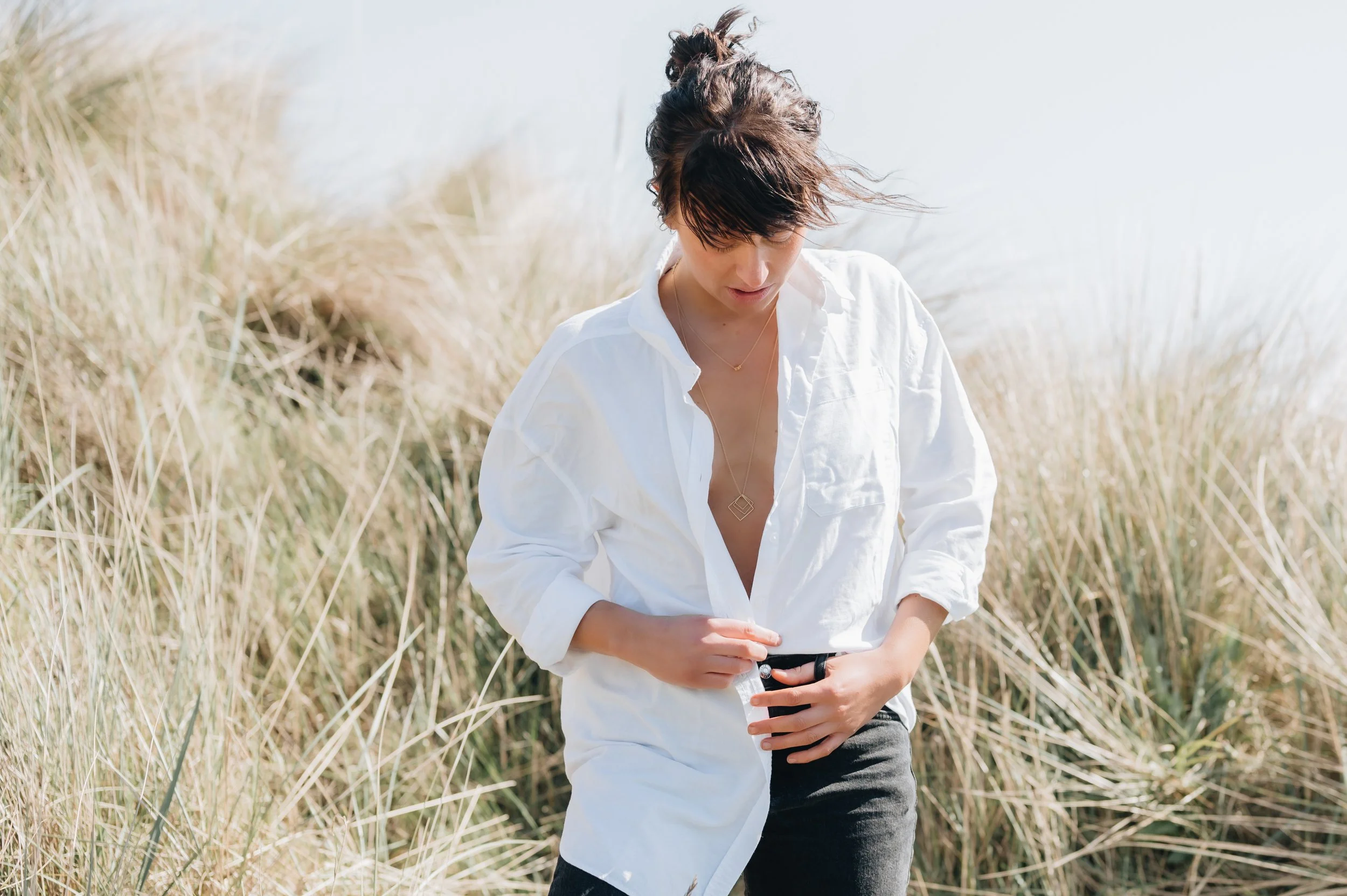 Woman in white shirt outdoors in a grassy field, adjusting her belt, with wind in her hair.