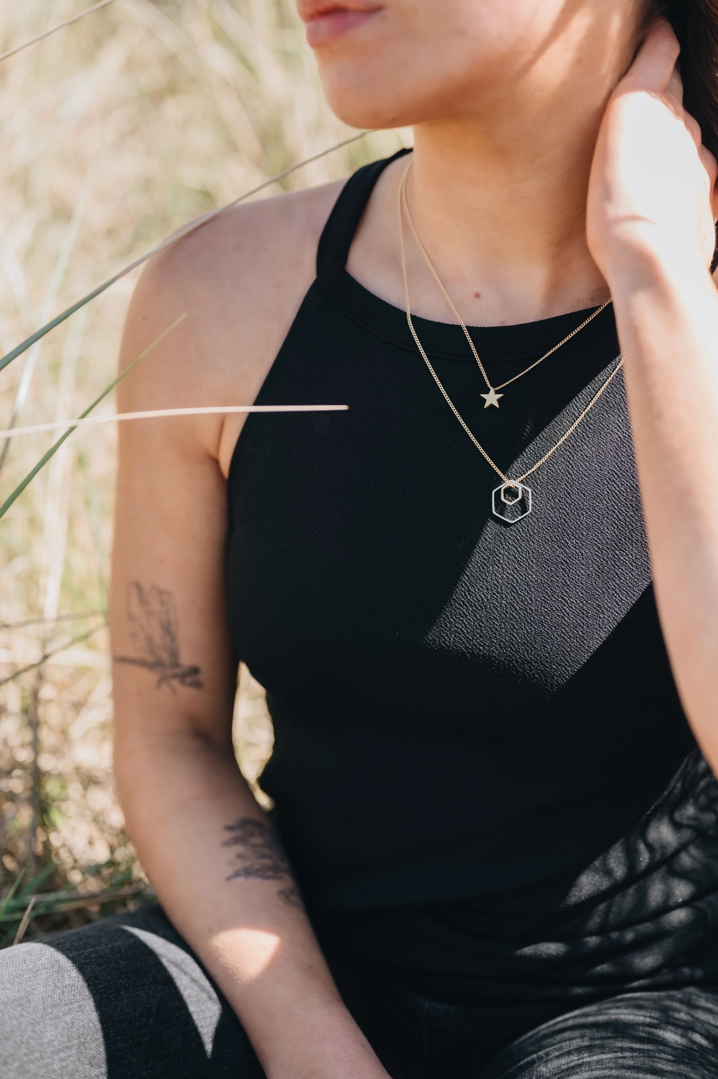 Close-up of a woman wearing a black sleeveless top with layered necklaces, including a star and hexagon pendant, sitting outdoors among tall grass.