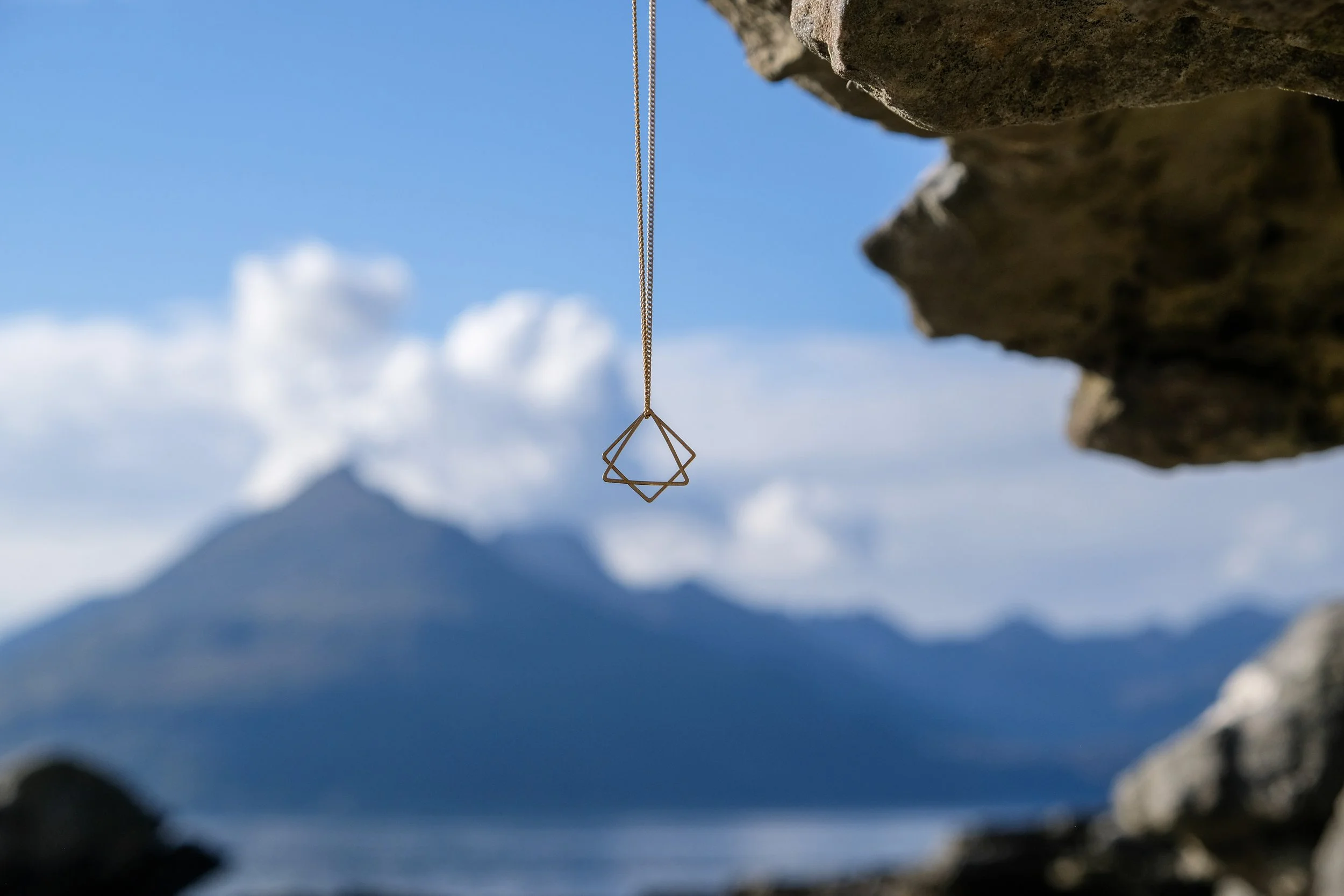 Necklace with geometric pendant hanging in front of a mountain and cloudy sky.