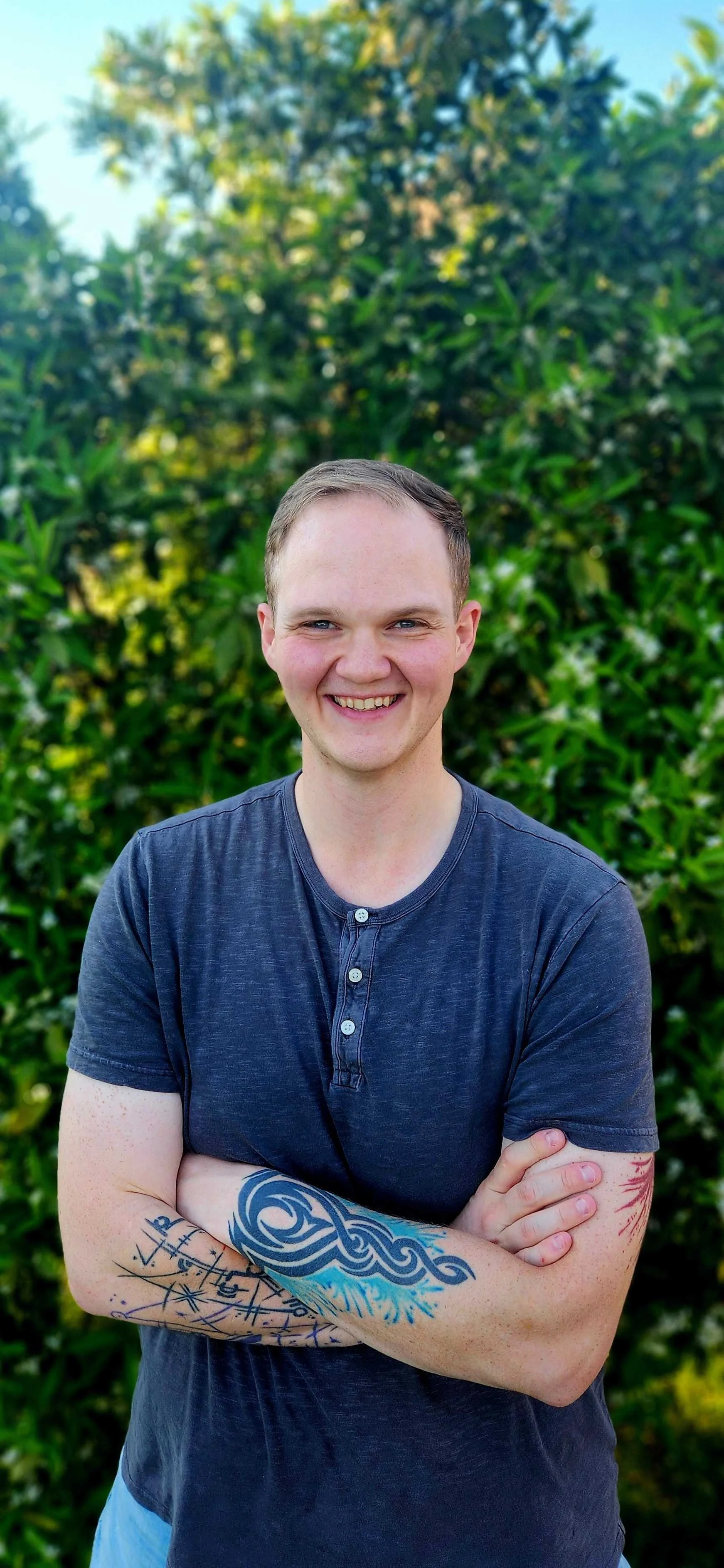 A young man with short light brown hair, smiling with arms crossed, standing outdoors in front of green bushes and a clear sky.