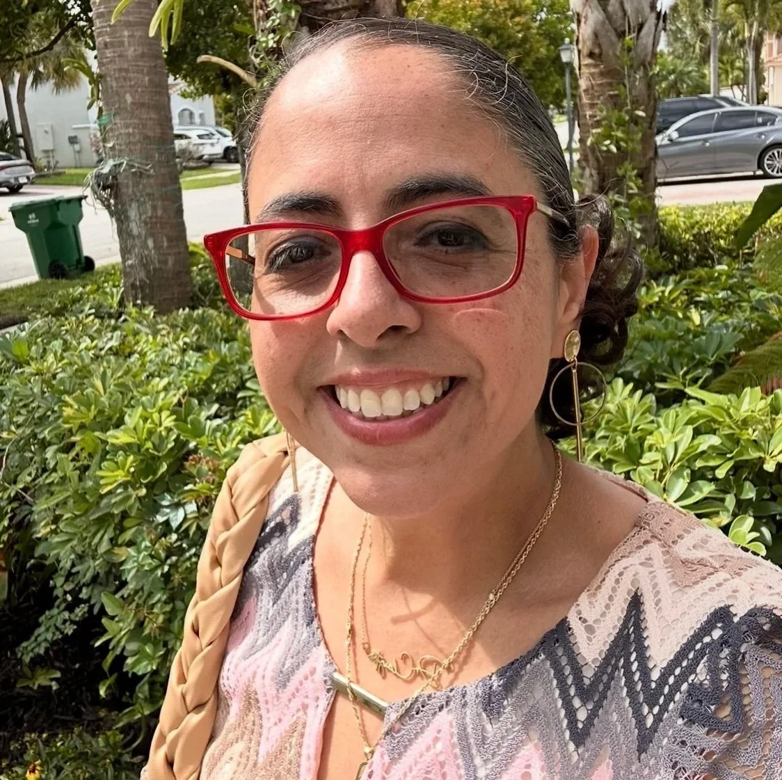 A woman smiling outdoors with trees and cars in the background. She is wearing red glasses, earrings, necklaces, and a patterned top.