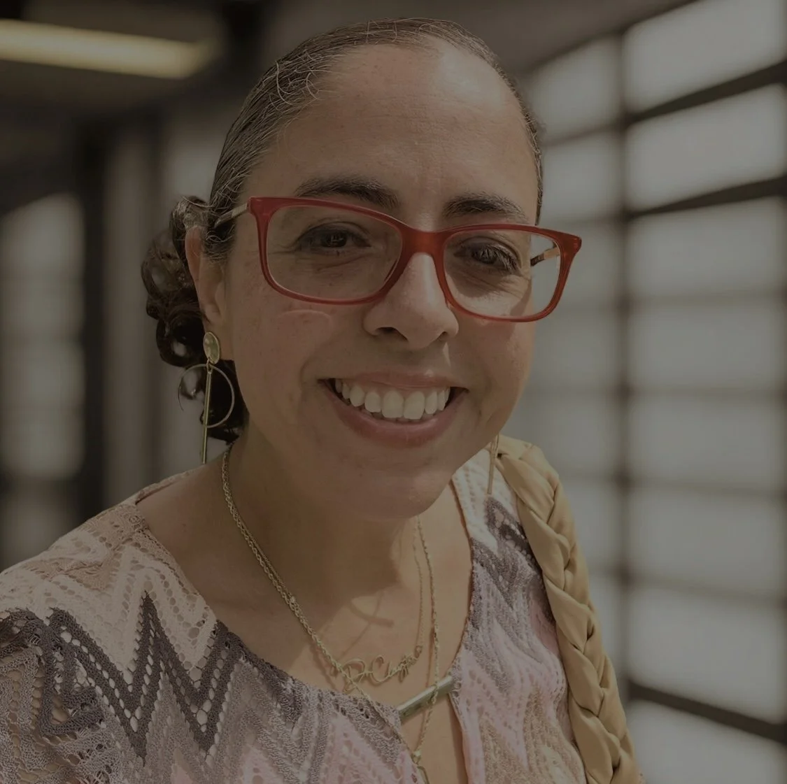 Close-up of a smiling woman with red glasses, braided hair, wearing earrings and layered necklaces, indoors near large windows.