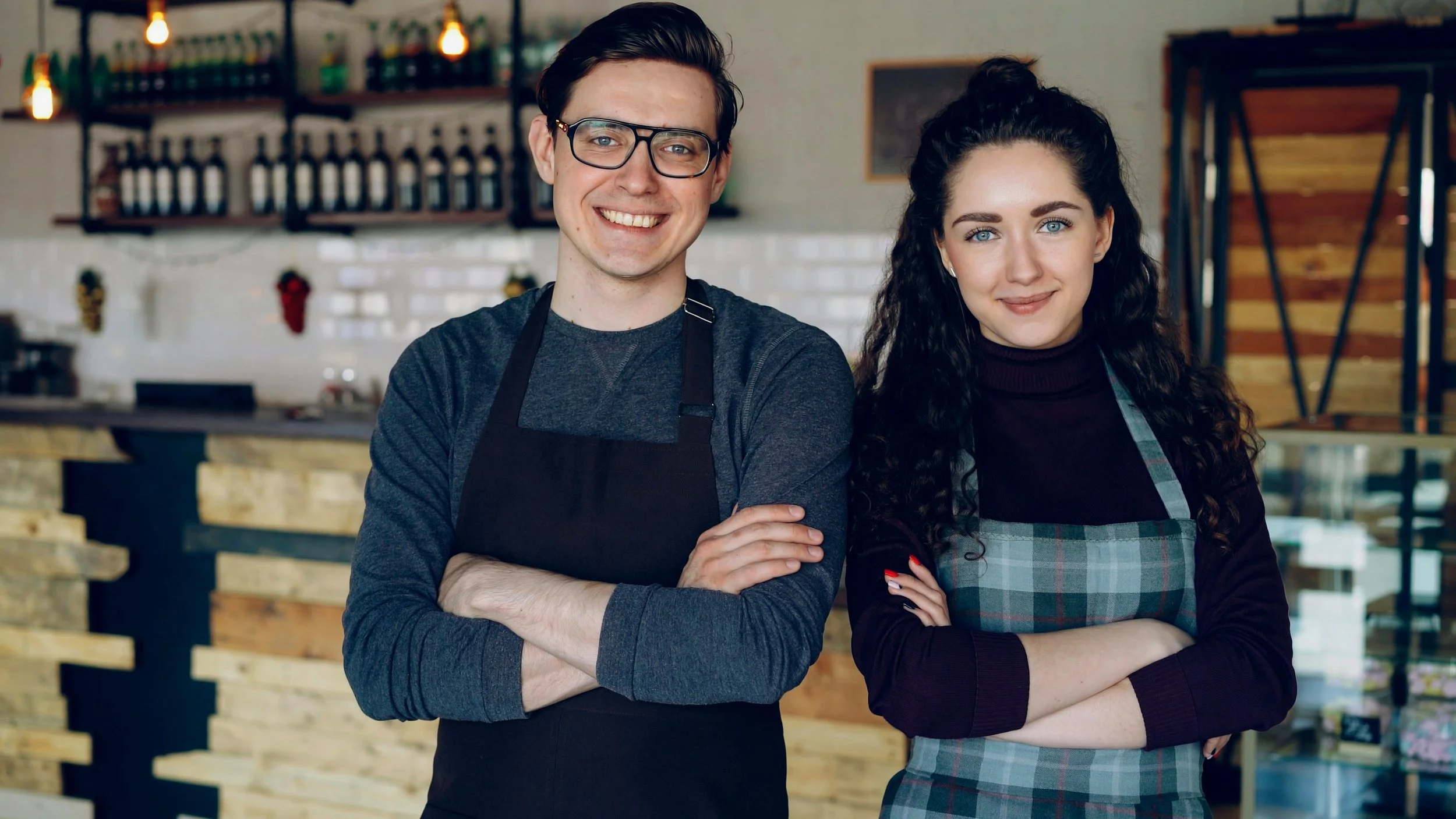 A smiling man and woman in aprons standing with arms crossed inside a cafe or restaurant with shelves of bottles and a counter behind them.