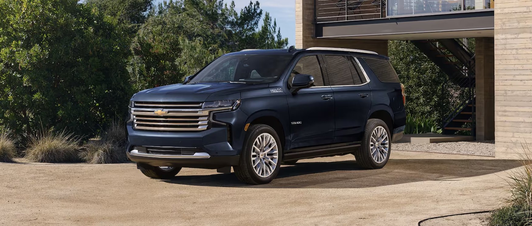 A dark blue Chevrolet SUV parked on a gravel driveway in front of a modern house with wooden exterior walls and an external staircase, surrounded by green bushes and trees.