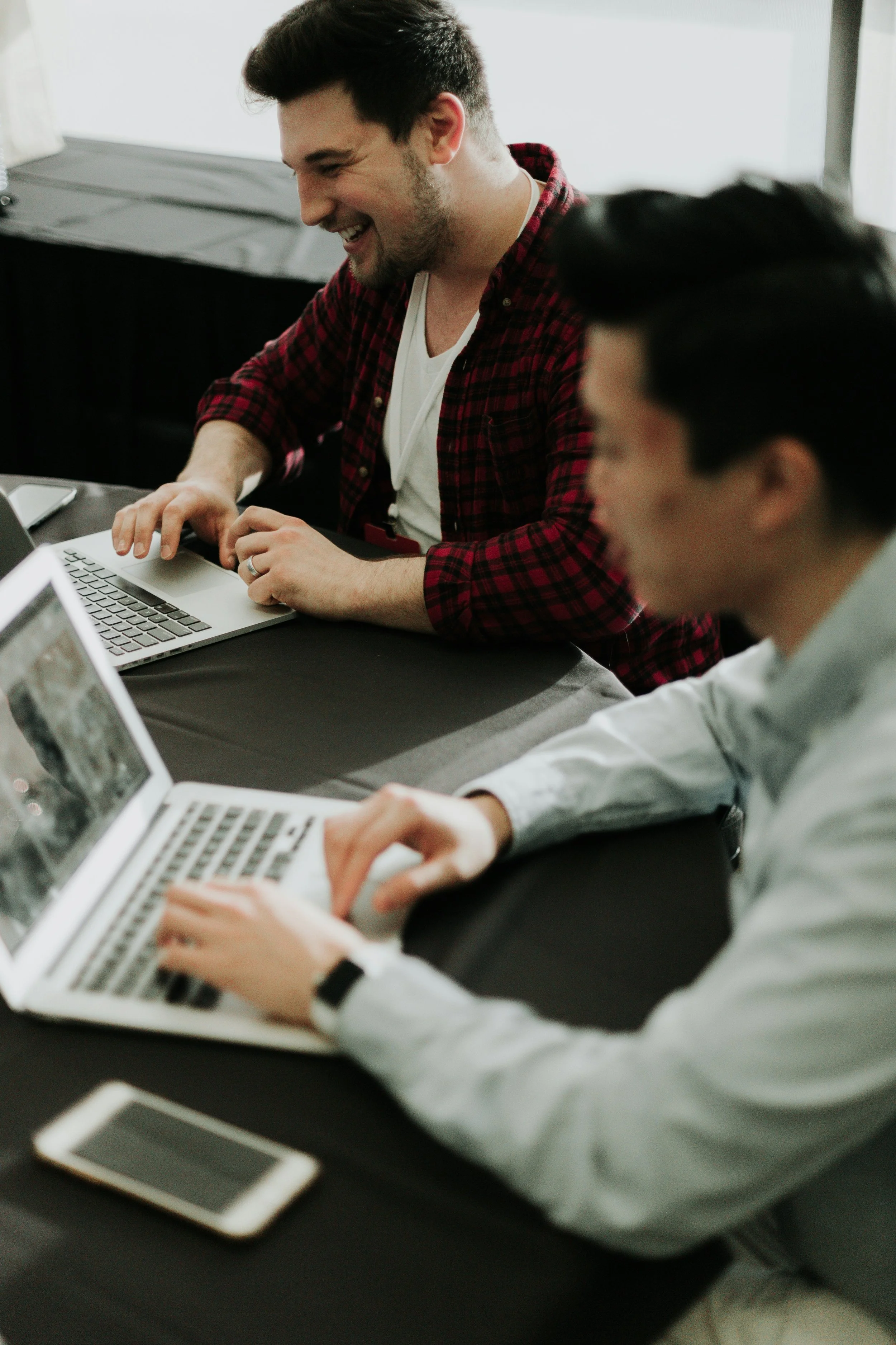 Two men sitting at a table using laptops, smiling and engaged in work or discussion, with a smartphone on the table.